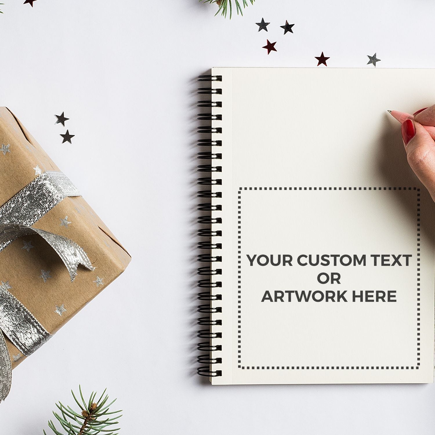 A hand writing in a notebook stamped with YOUR CUSTOM TEXT OR ARTWORK HERE using a Custom Rubber Stamp Size 6 x 6, next to a gift box.