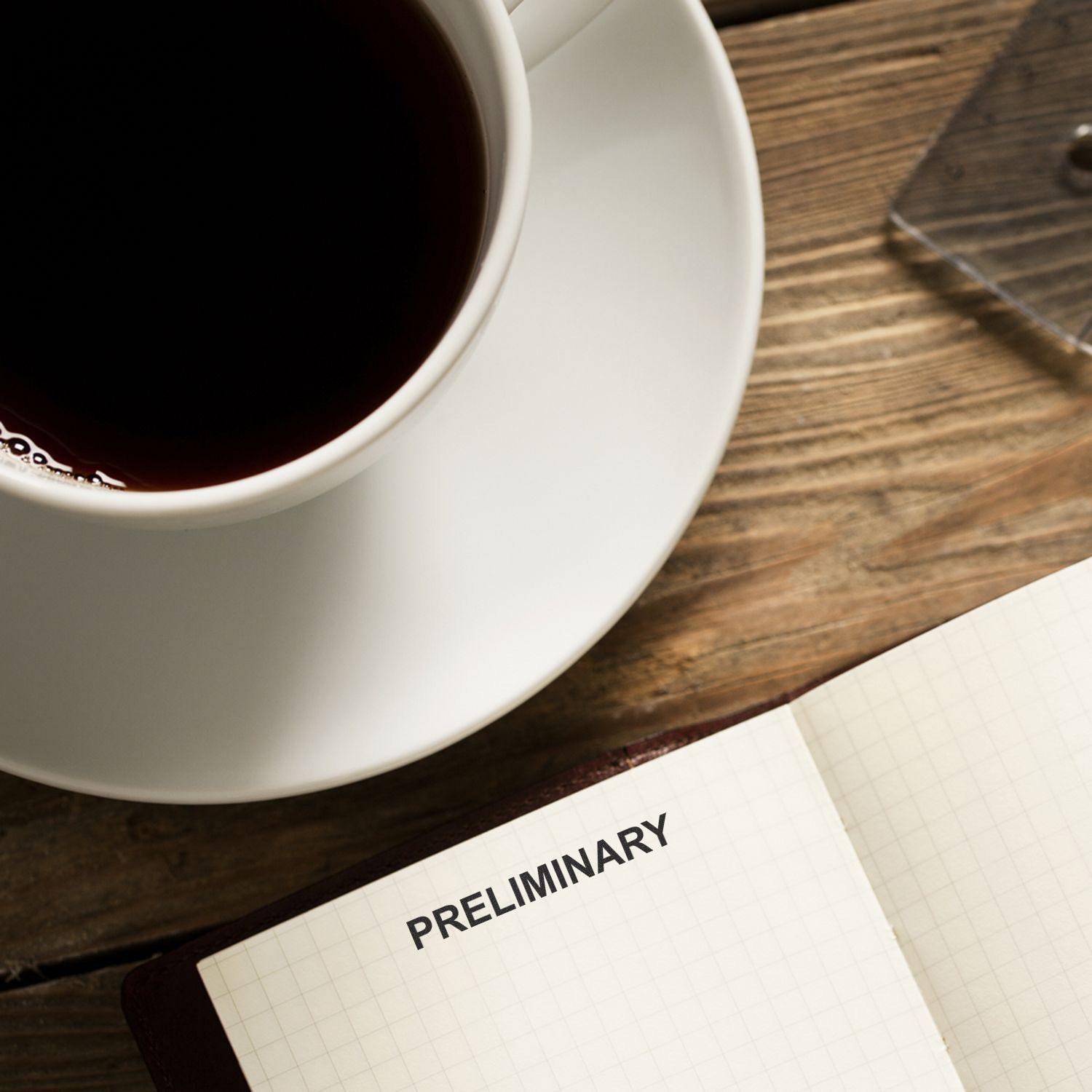 Self Inking Preliminary Stamp marking PRELIMINARY on a notebook page, next to a cup of coffee on a wooden table.