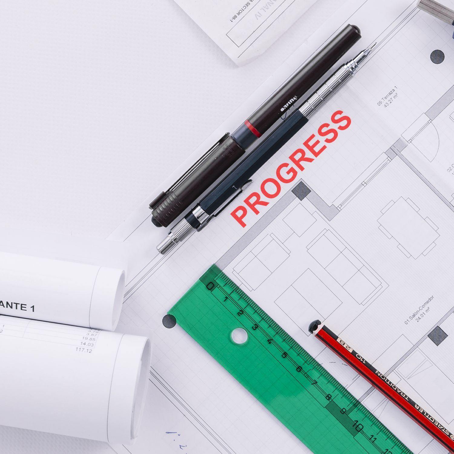 Slim Pre-Inked Progress Stamp used on architectural plans with pens, ruler, and rolled blueprints on a white surface.