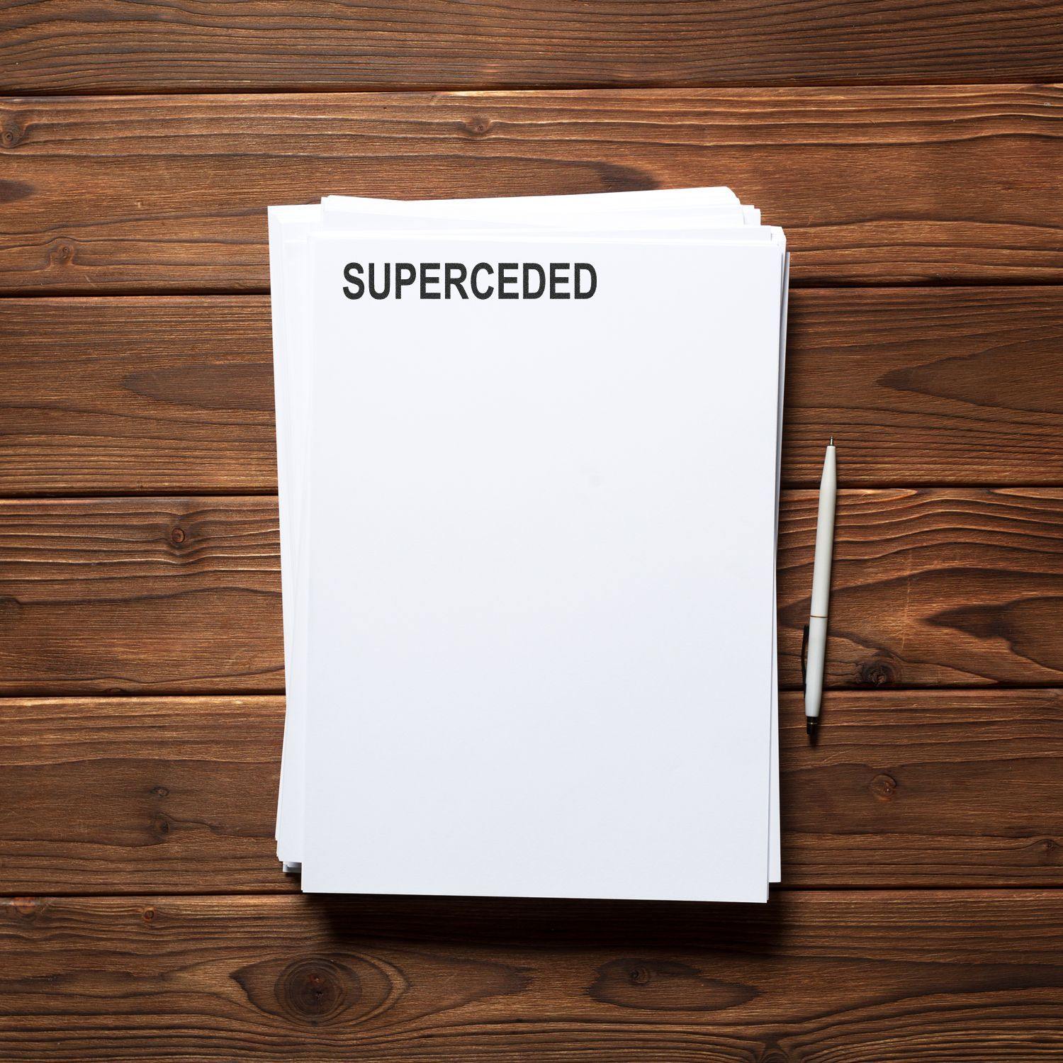 Slim Pre-Inked Superceded Stamp marking SUPERCEDED on a stack of white papers, with a pen placed beside the papers on a wooden surface.