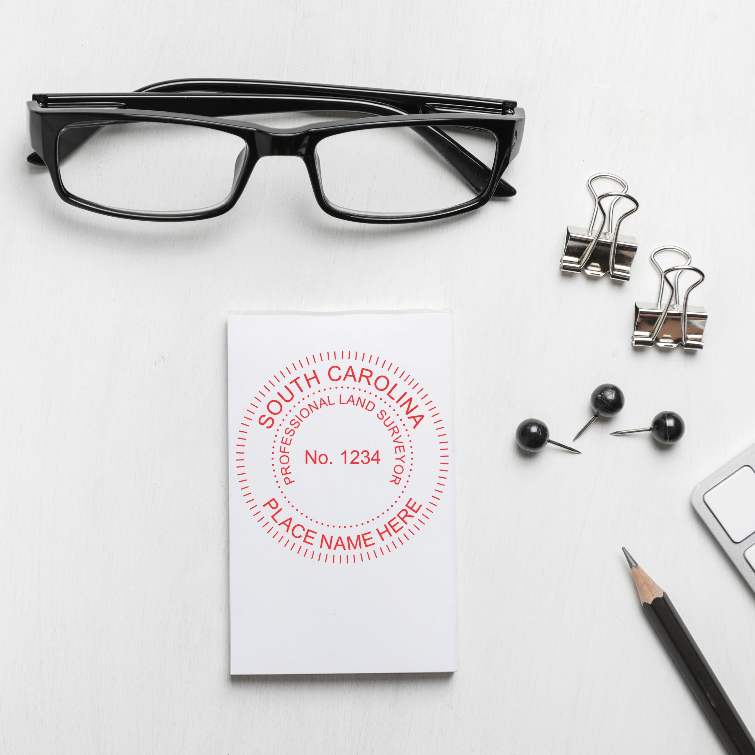 Self Inking South Carolina Land Surveyor Stamp on paper, surrounded by glasses, binder clips, push pins, pencil, and keyboard.