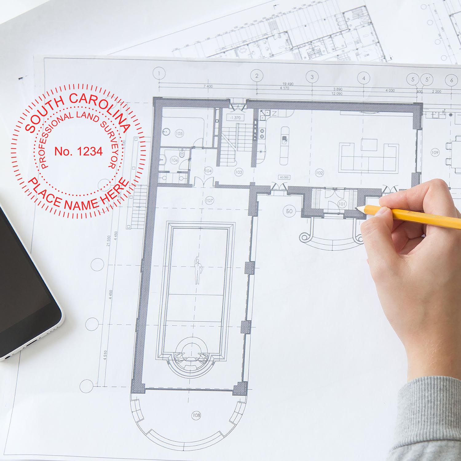 Person using Premium MaxLight Pre-Inked South Carolina Surveyors Stamp on architectural plans with a pencil and smartphone nearby.