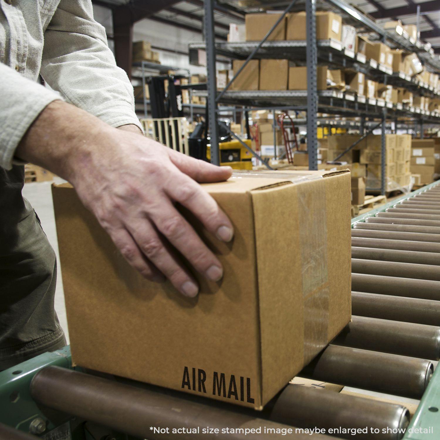 A person using a Slim Pre-Inked Air Mail Stamp on a cardboard box in a warehouse with shelves of boxes in the background.