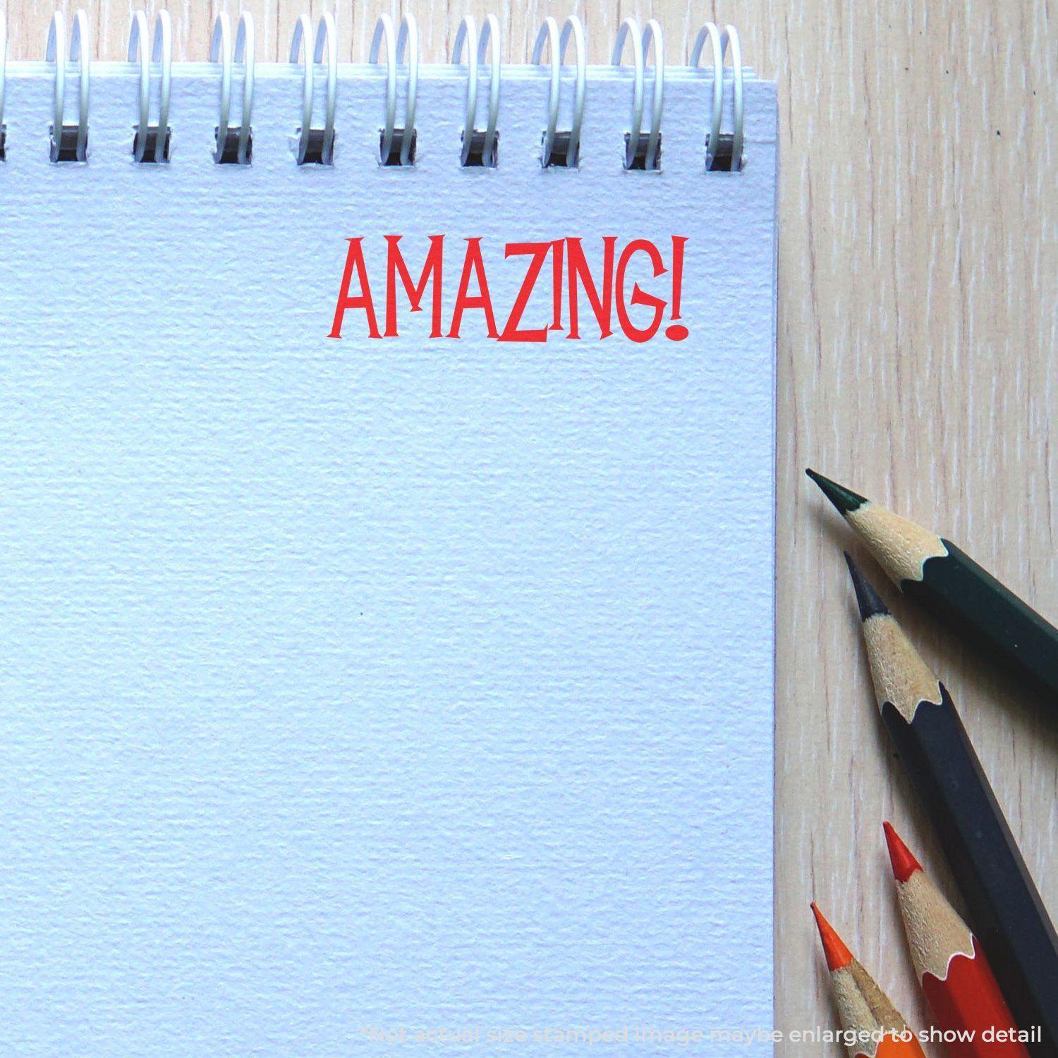 Notebook with the word 'AMAZING!' stamped in red using an Amazing Rubber Stamp, next to colored pencils on a wooden surface.