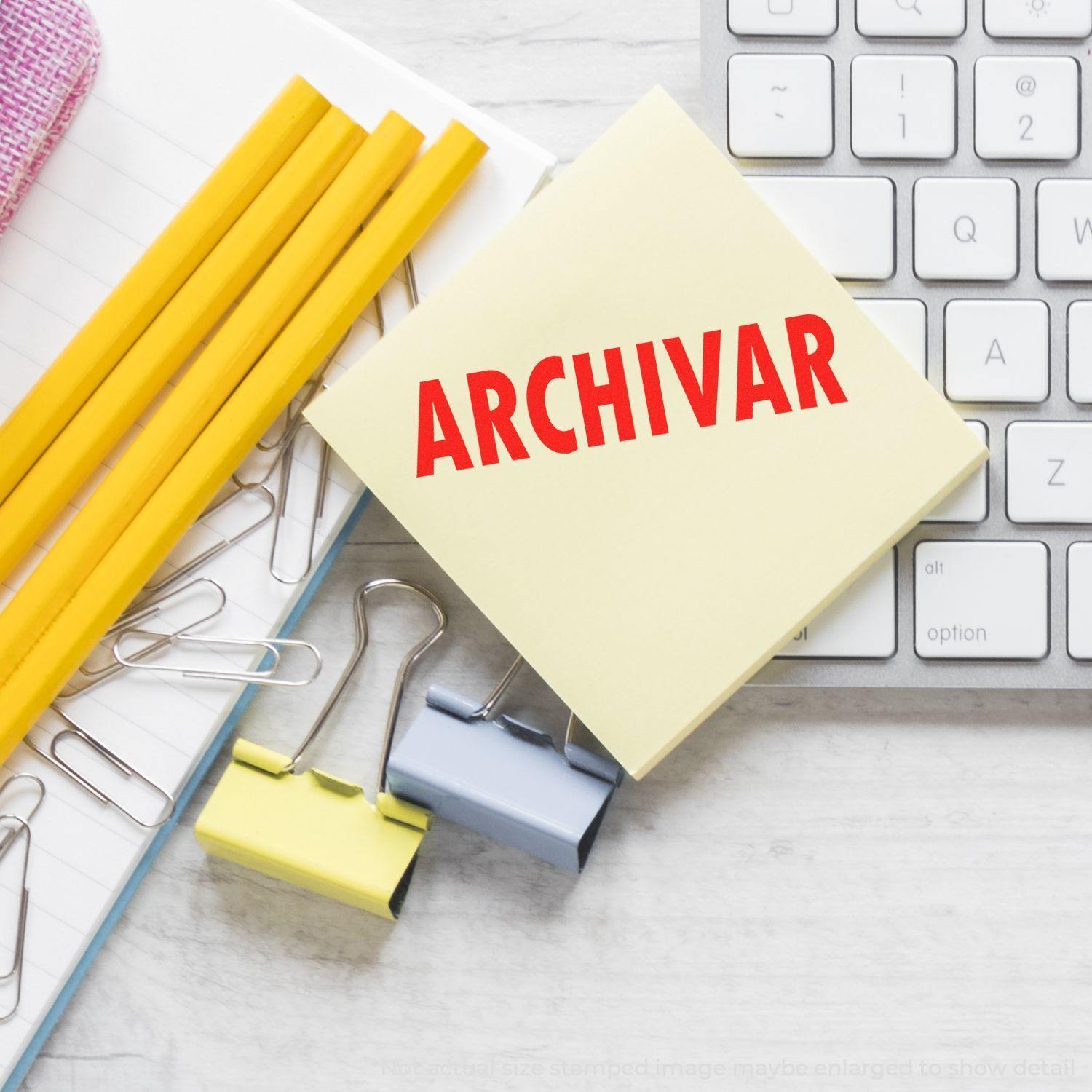 Large Self Inking Archivar Stamp on a yellow sticky note beside pencils, paper clips, and a keyboard on a desk.