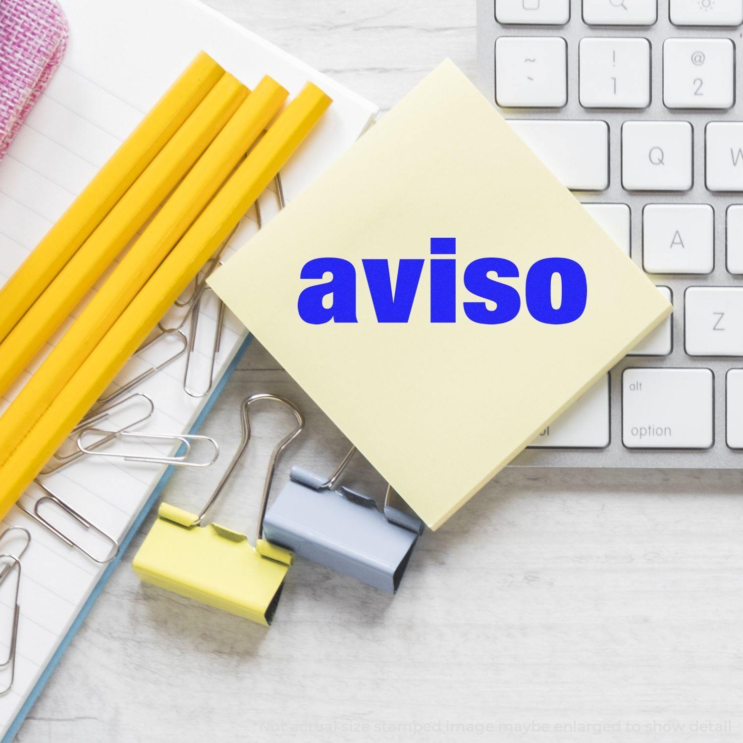 Large Self Inking Aviso Stamp on a yellow sticky note beside a keyboard, pencils, paper clips, and binder clips on a desk.