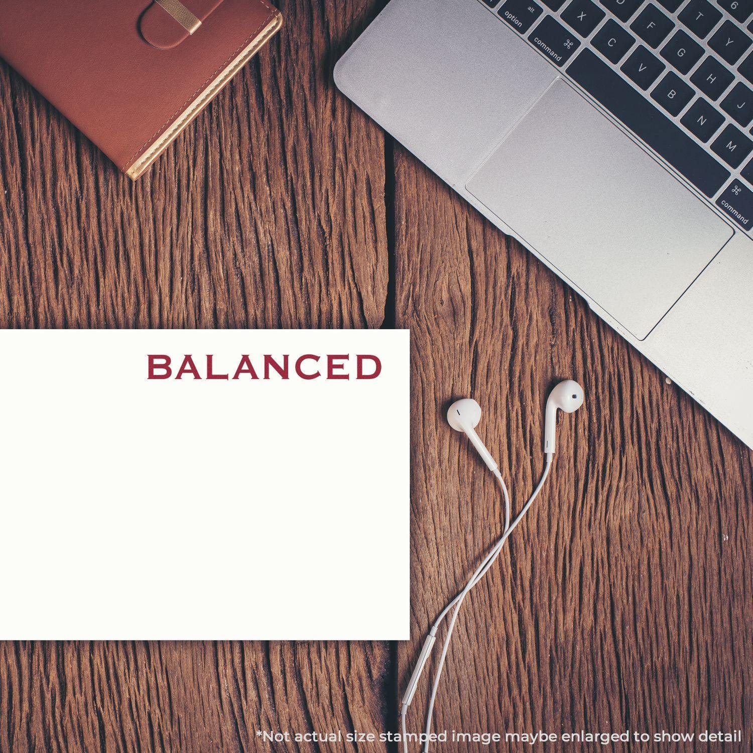 Slim Pre-Inked Balanced Stamp on white paper next to a laptop, notebook, and earphones on a wooden desk.