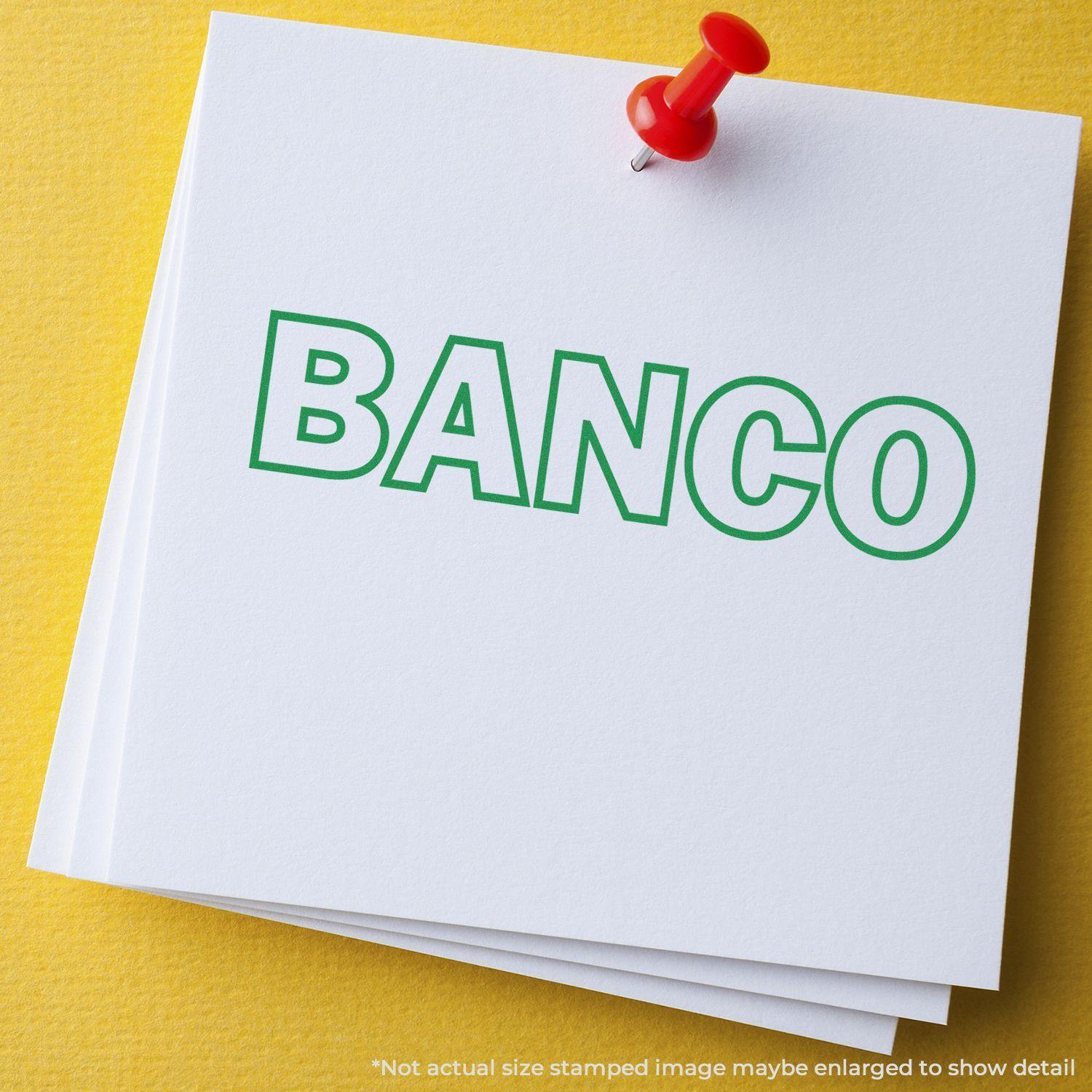 White paper with green 'BANCO' text stamped using a Slim Pre-Inked Banco Stamp, pinned with a red pushpin on a yellow surface.