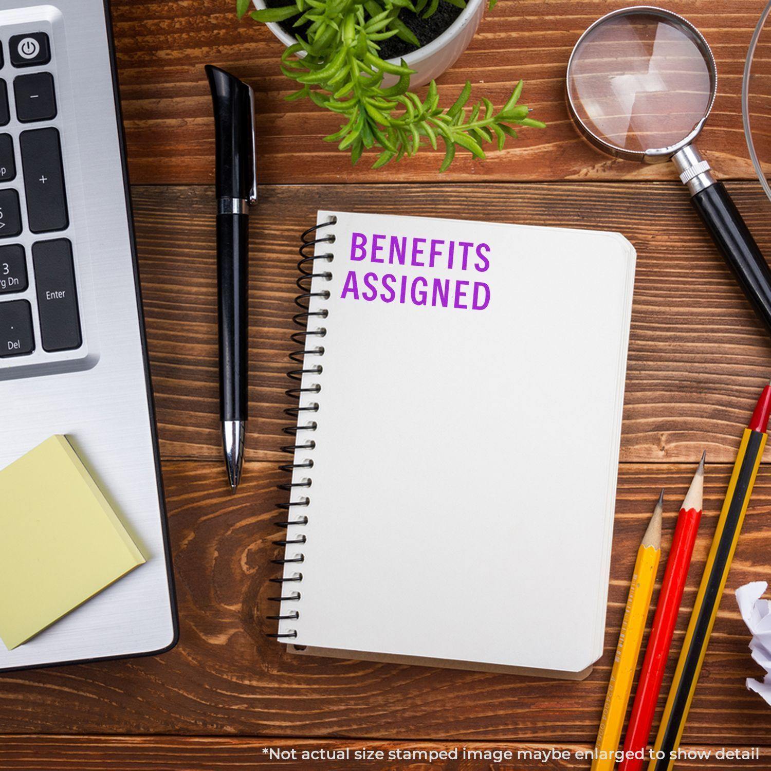 A notebook with Benefits Assigned rubber stamp on a wooden desk, surrounded by a laptop, pen, sticky notes, and stationery.