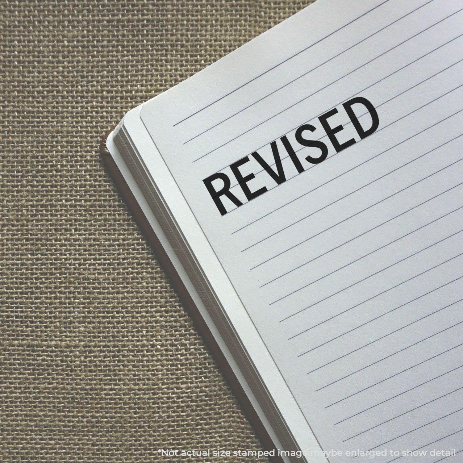 Large Pre-Inked Revised Stamp in use, marking the word REVISED on a lined notebook page placed on a textured surface.