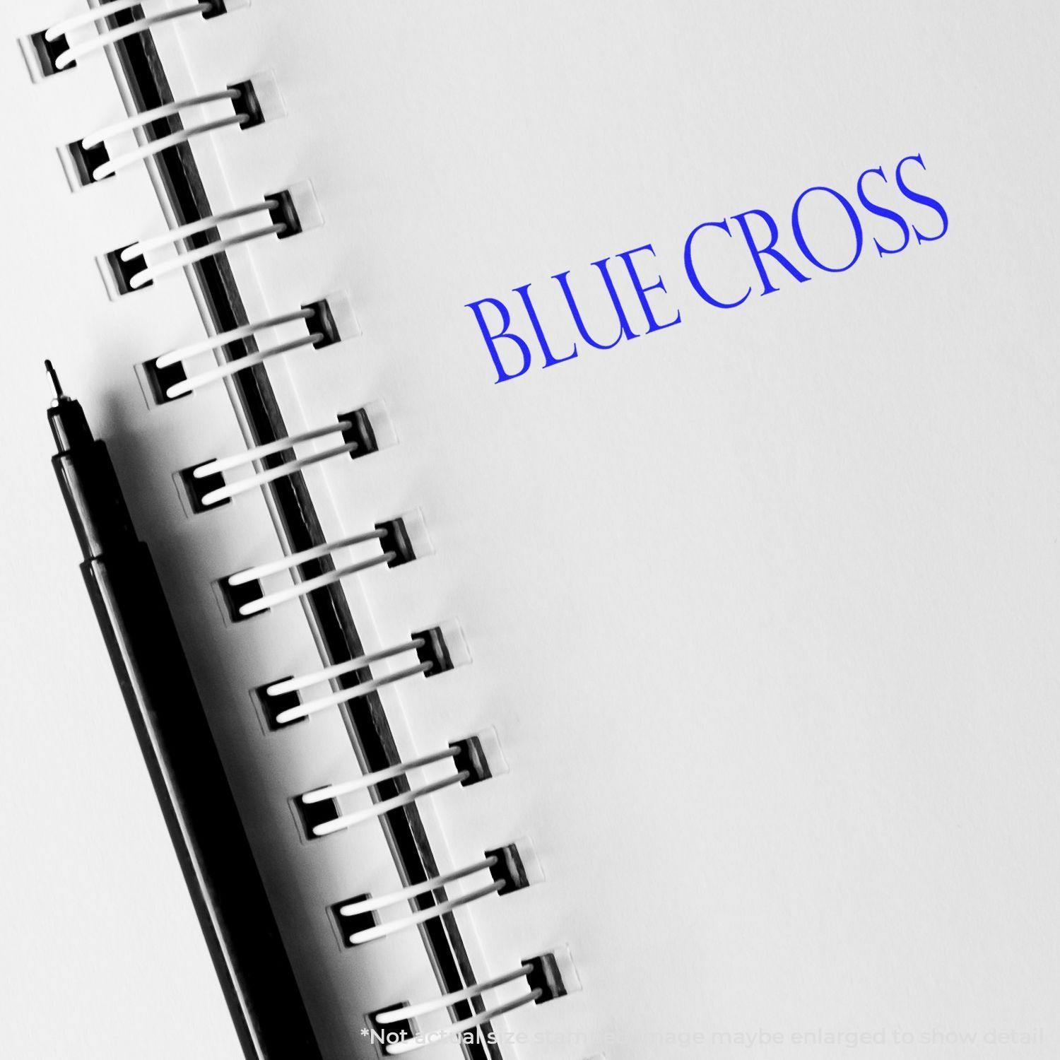 Slim Pre-Inked Blue Shield Stamp imprinting BLUE CROSS on a white notebook page with a black pen beside it.