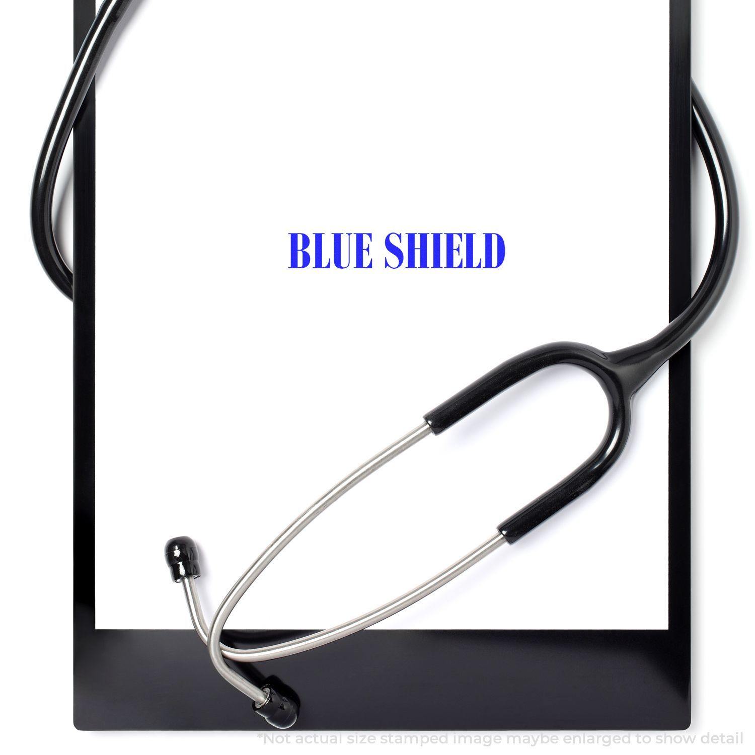A self-inking stamp with a stamped image showing how the text "BLUE SHIELD" in a large bold font is displayed by it.