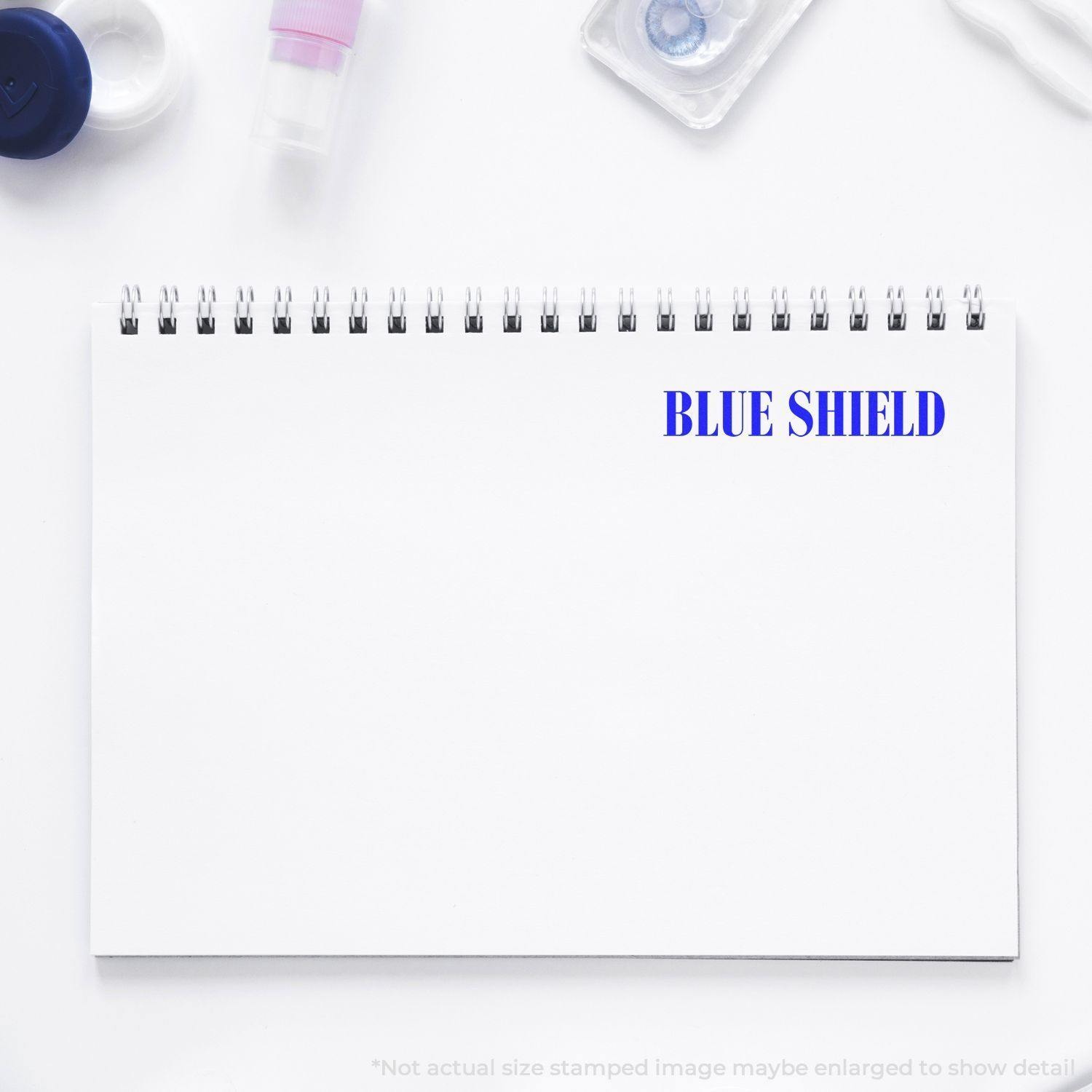 Blue Shield rubber stamp impression on a white spiral notebook, surrounded by office supplies.