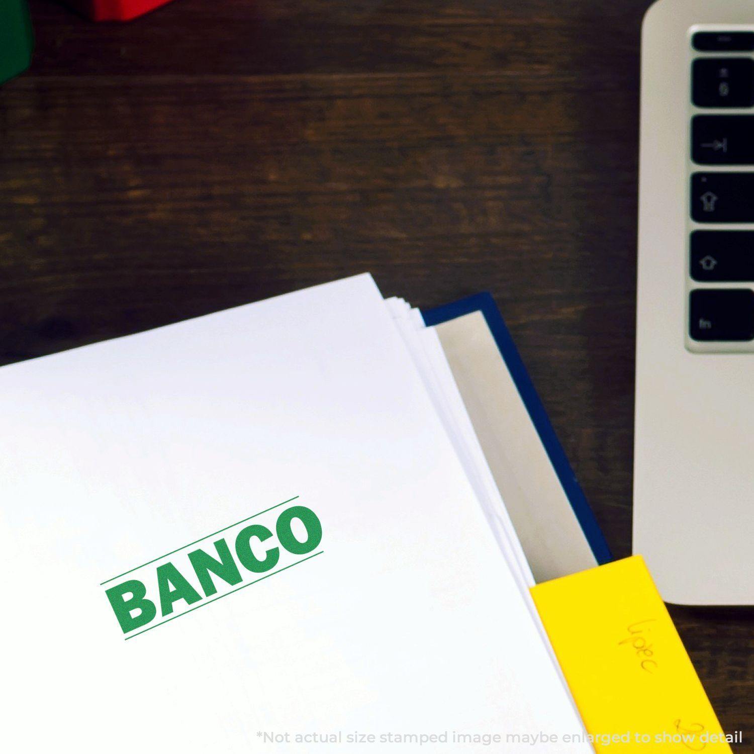 A document stamped with BANCO using the Large Self Inking Bold Banco Stamp, placed on a wooden desk next to a laptop and yellow sticky notes.