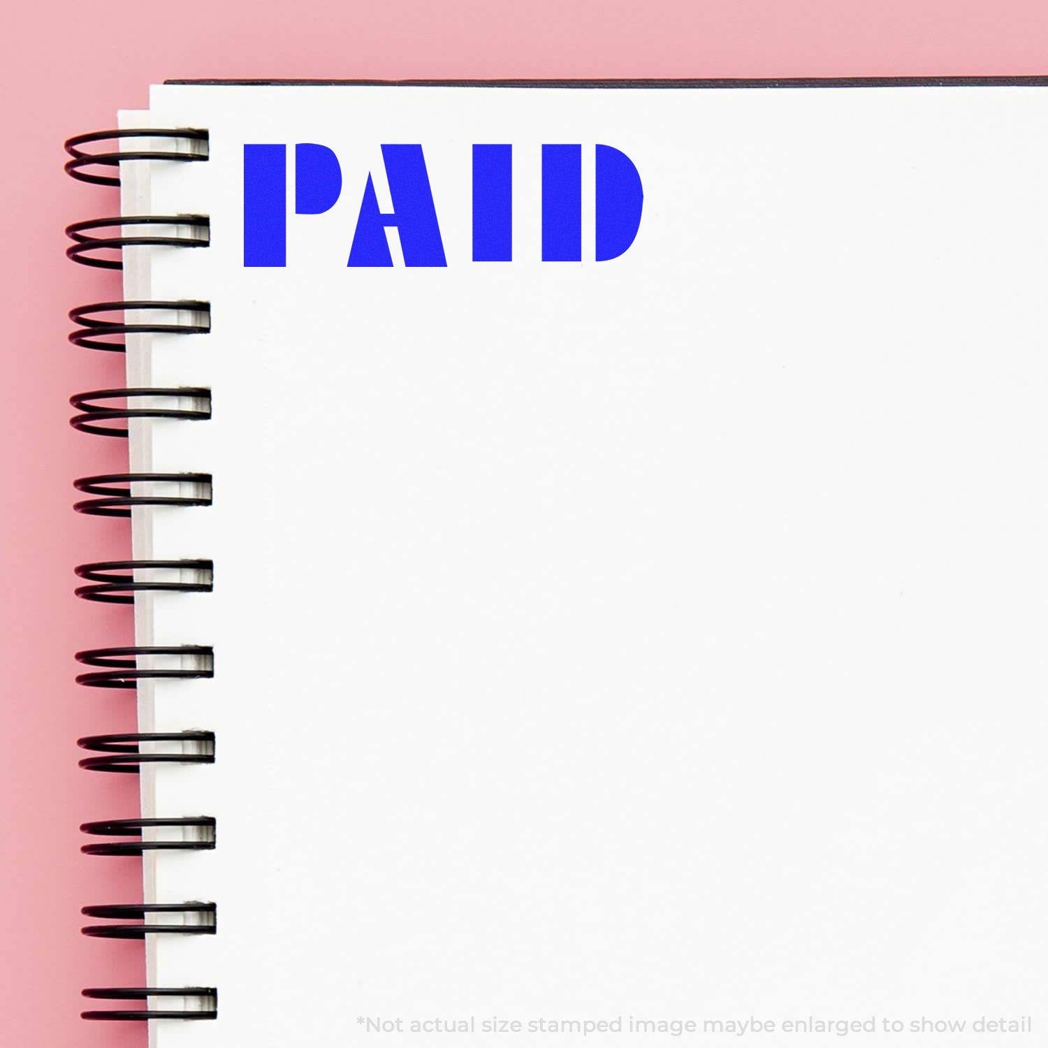 Slim Pre-Inked Bold Paid Stamp in blue ink on a white spiral notebook with a pink background.