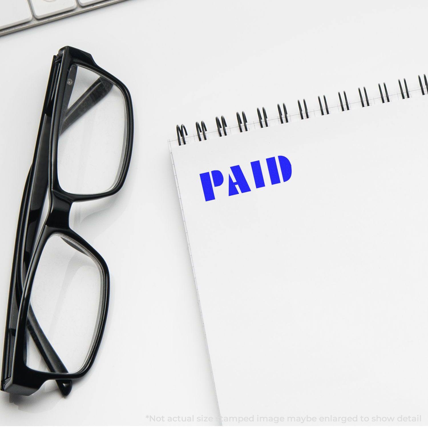 Large Bold Paid Rubber Stamp in blue ink on a white notepad, with black glasses and a keyboard in the background.