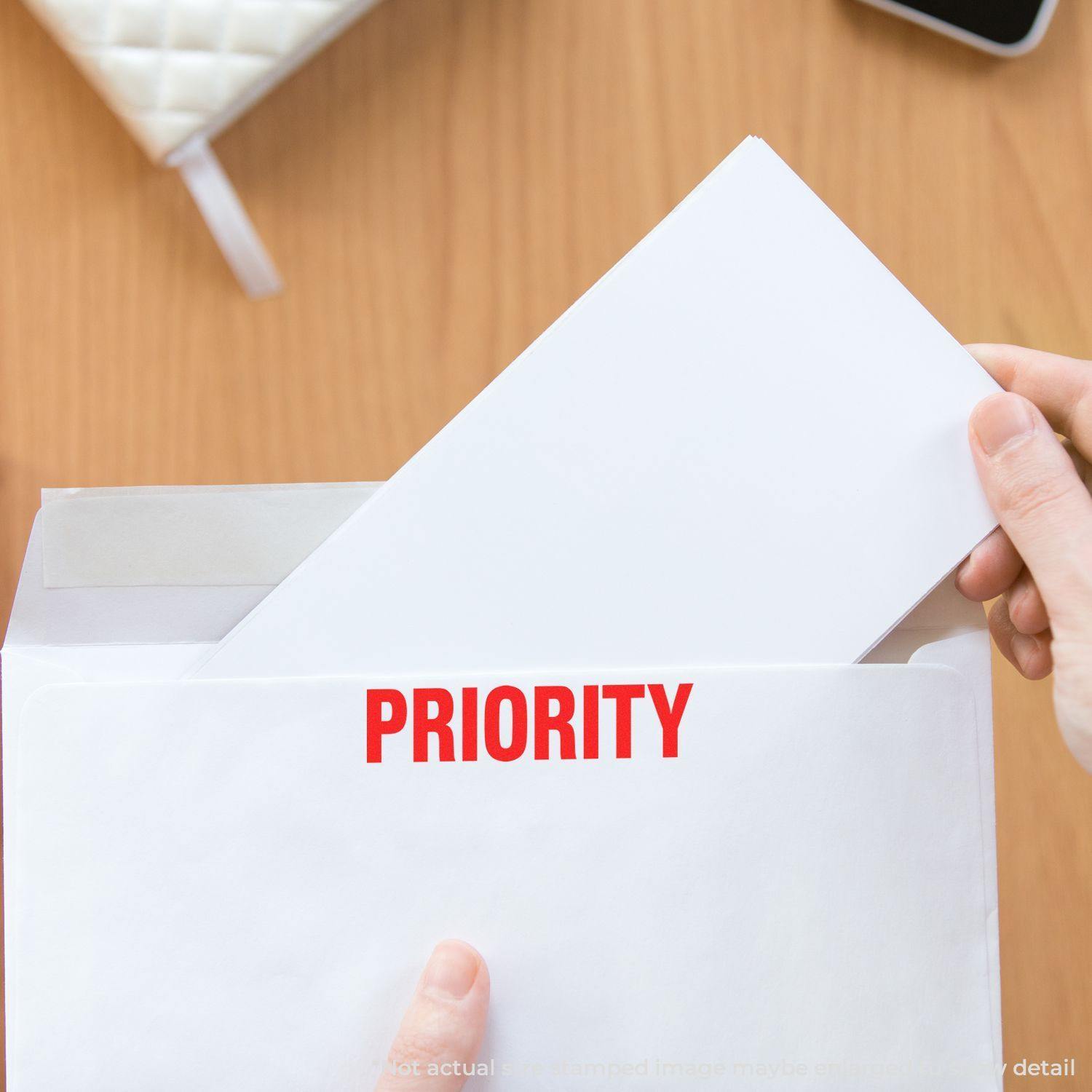 A hand placing an envelope stamped with PRIORITY in bold red letters into a larger envelope on a wooden table.