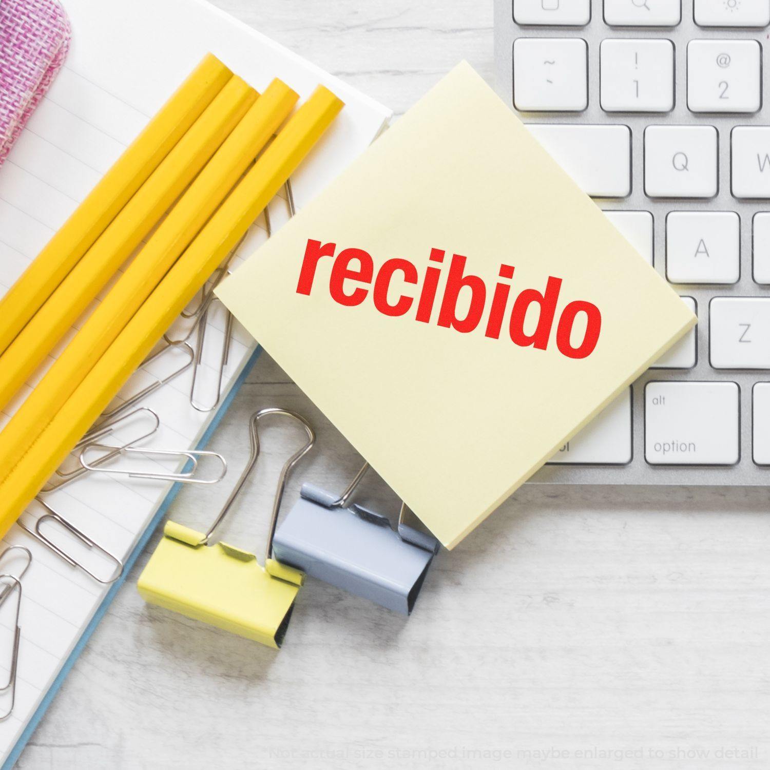 Large Self Inking Bold Recibido Stamp on a yellow sticky note, surrounded by pencils, paper clips, and a keyboard on a desk.