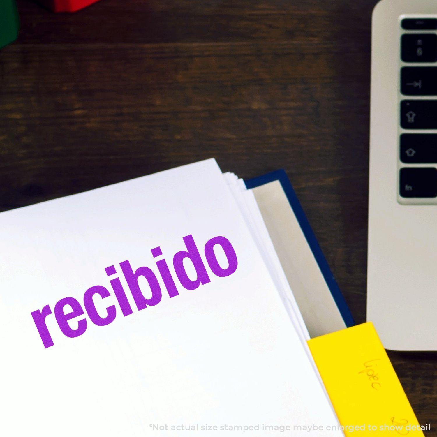 Bold Recibido Rubber Stamp in use, stamping recibido in purple ink on a white document, next to a laptop on a wooden desk.