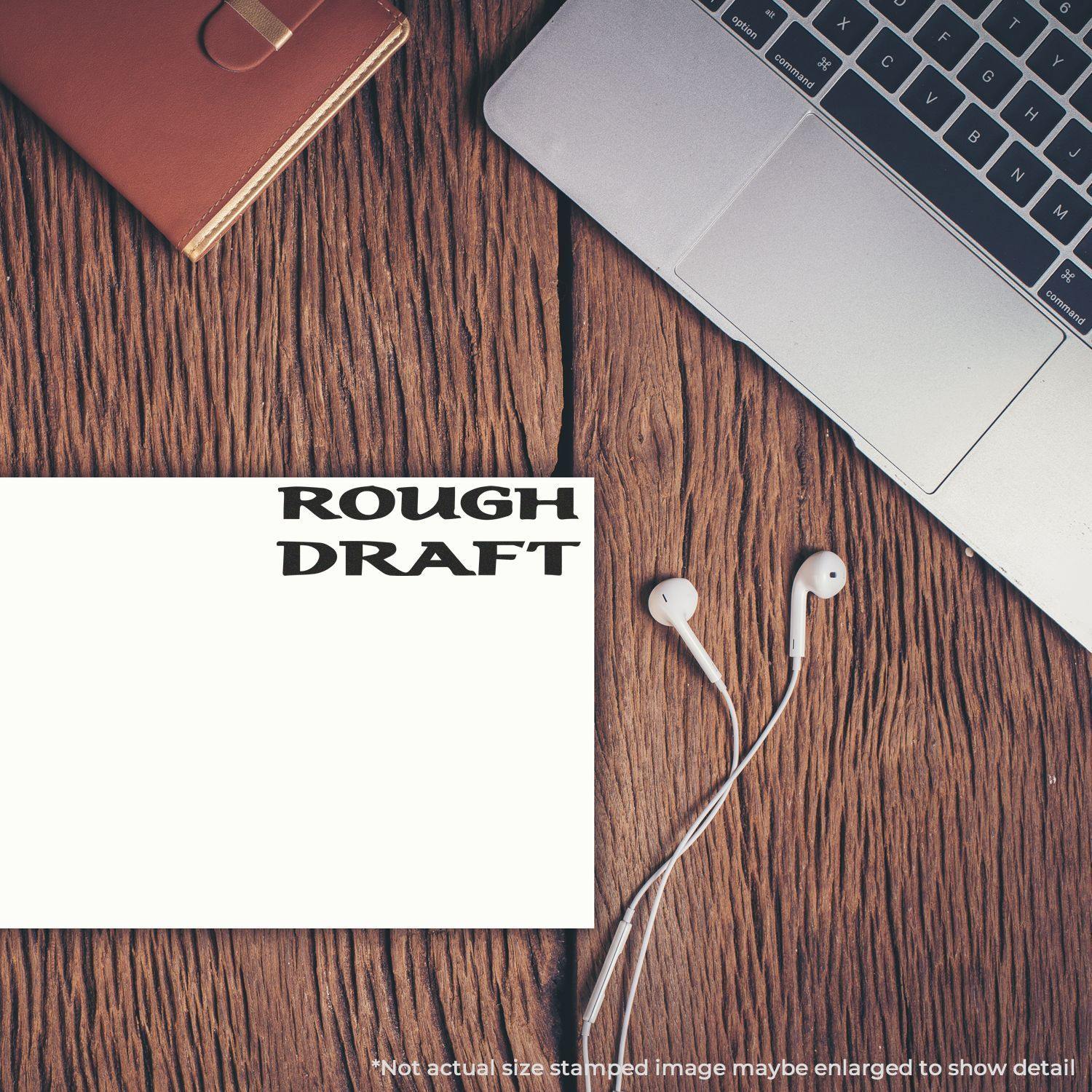 Large Self Inking Bold Rough Draft Stamp used on paper next to a laptop, notebook, and earphones on a wooden desk.