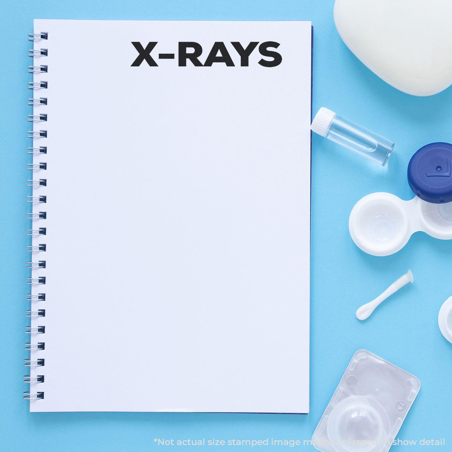 Large Self Inking Bold X-Rays Stamp used on a white spiral notebook, surrounded by medical items on a blue background.