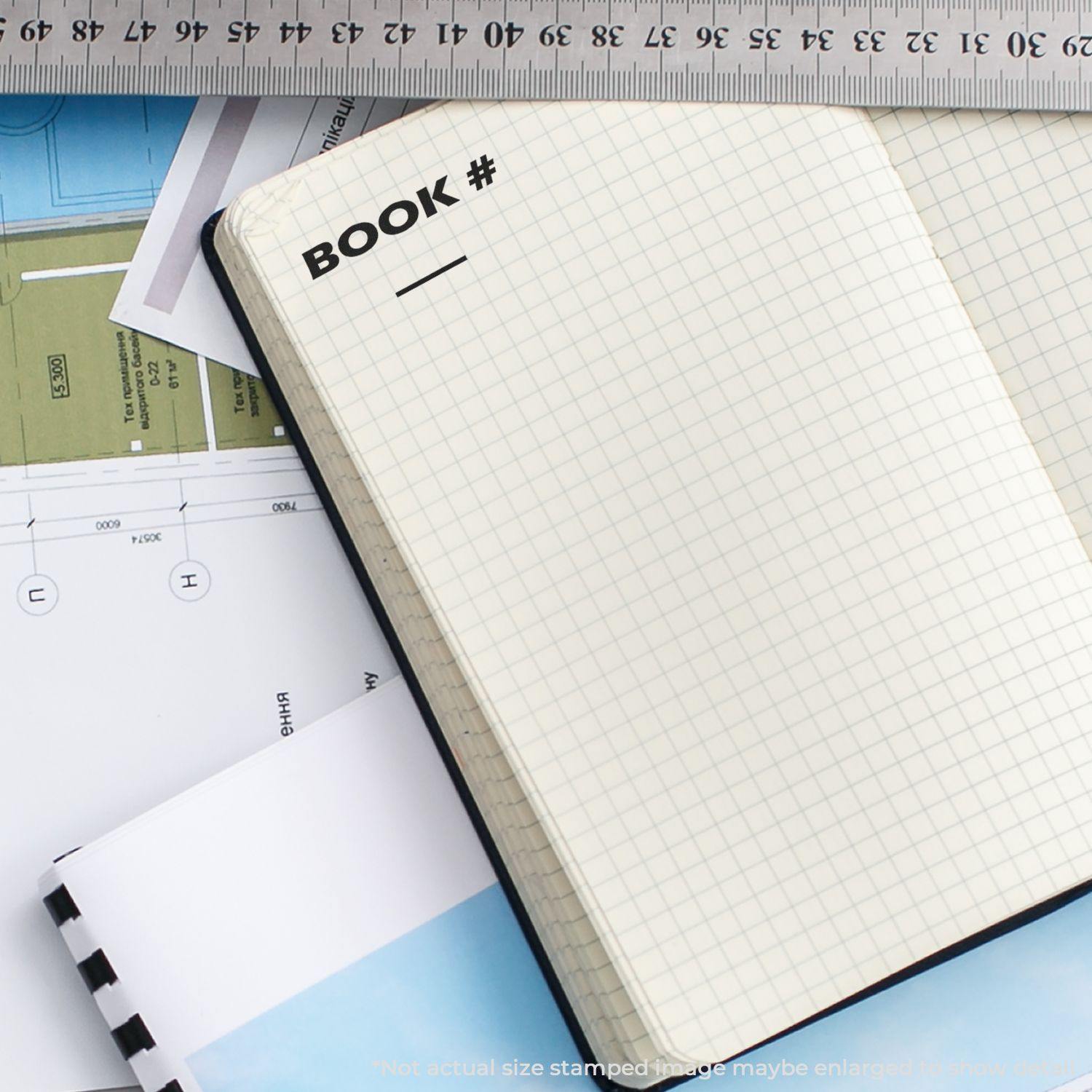 Open notebook stamped with BOOK # using the Book Rubber Stamp, placed on a desk with a ruler and architectural plans.