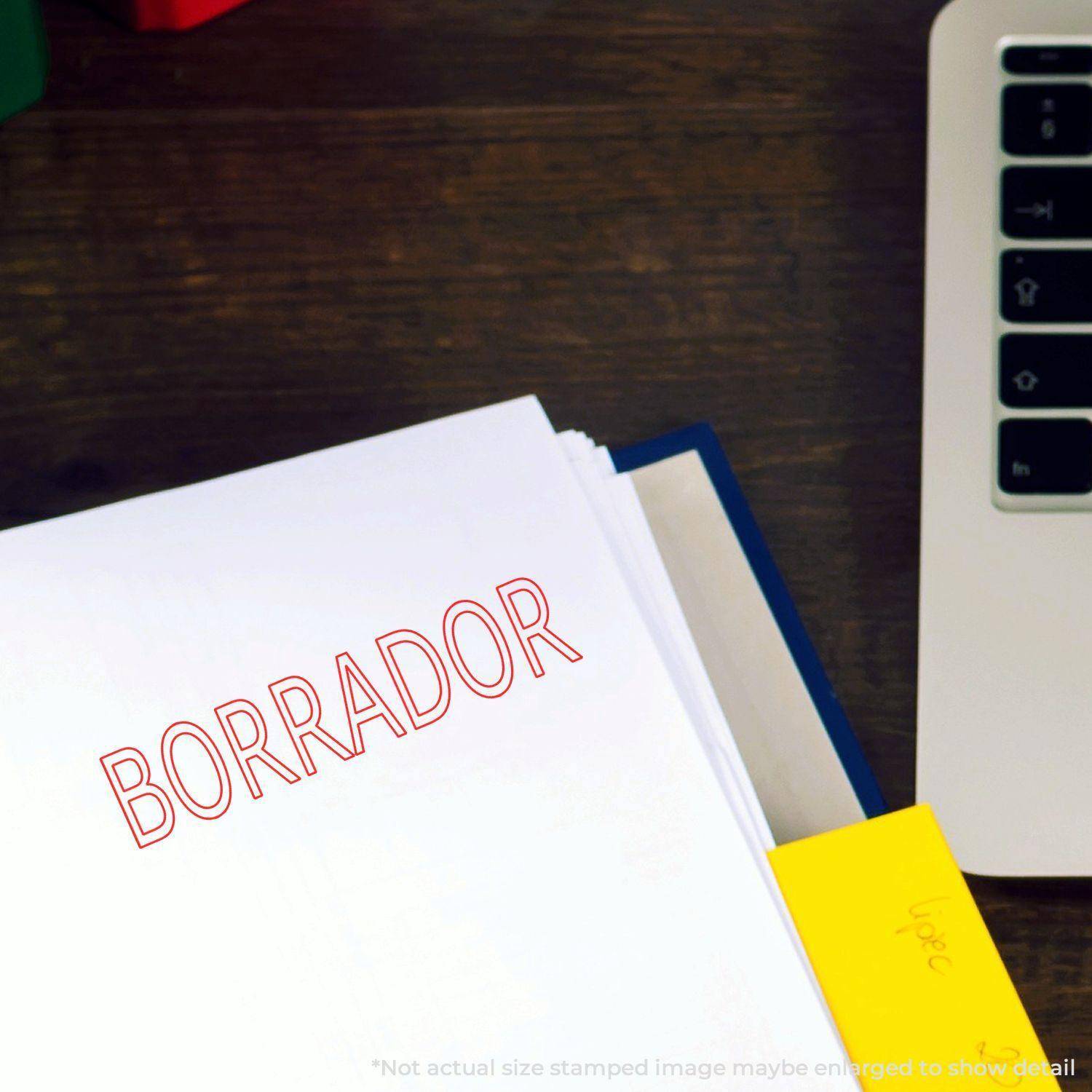 Large Self Inking Borrador Stamp in red ink on a white paper, next to a laptop and a yellow sticky note on a wooden desk.