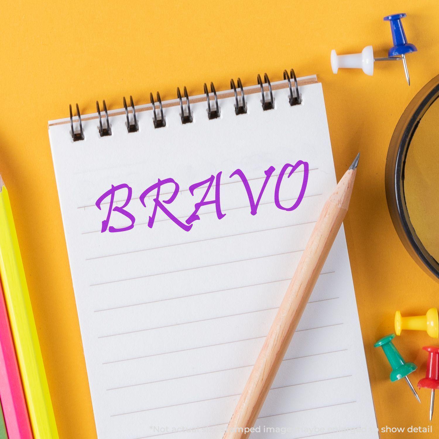 A Large Pre-Inked Bravo Stamp is used on a spiral notebook, surrounded by colorful pencils, push pins, and a pencil on a yellow background.