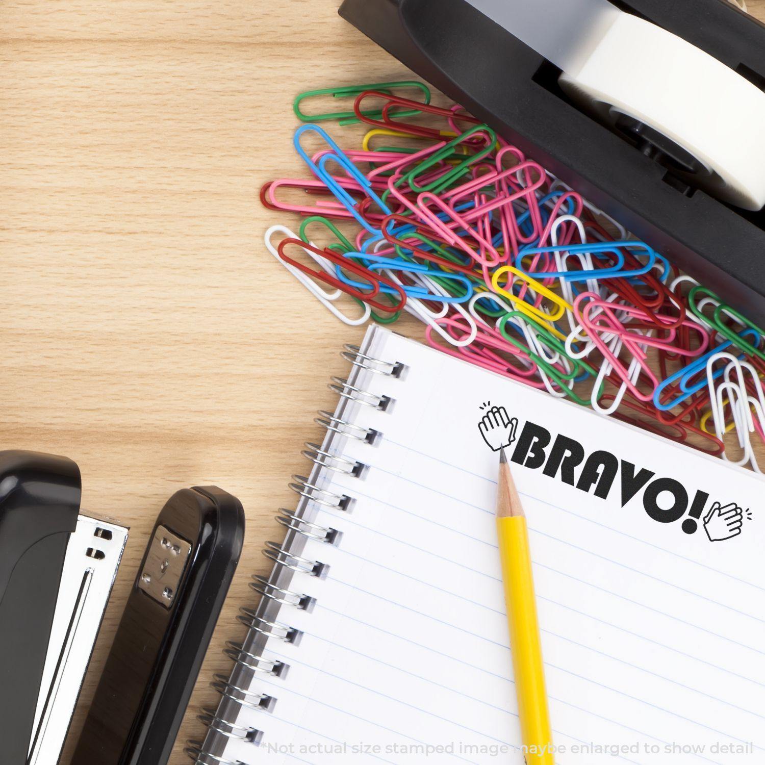 Large Pre-Inked Bravo with Hands Stamp on a notebook, surrounded by colorful paper clips, a stapler, and a tape dispenser on a wooden desk.