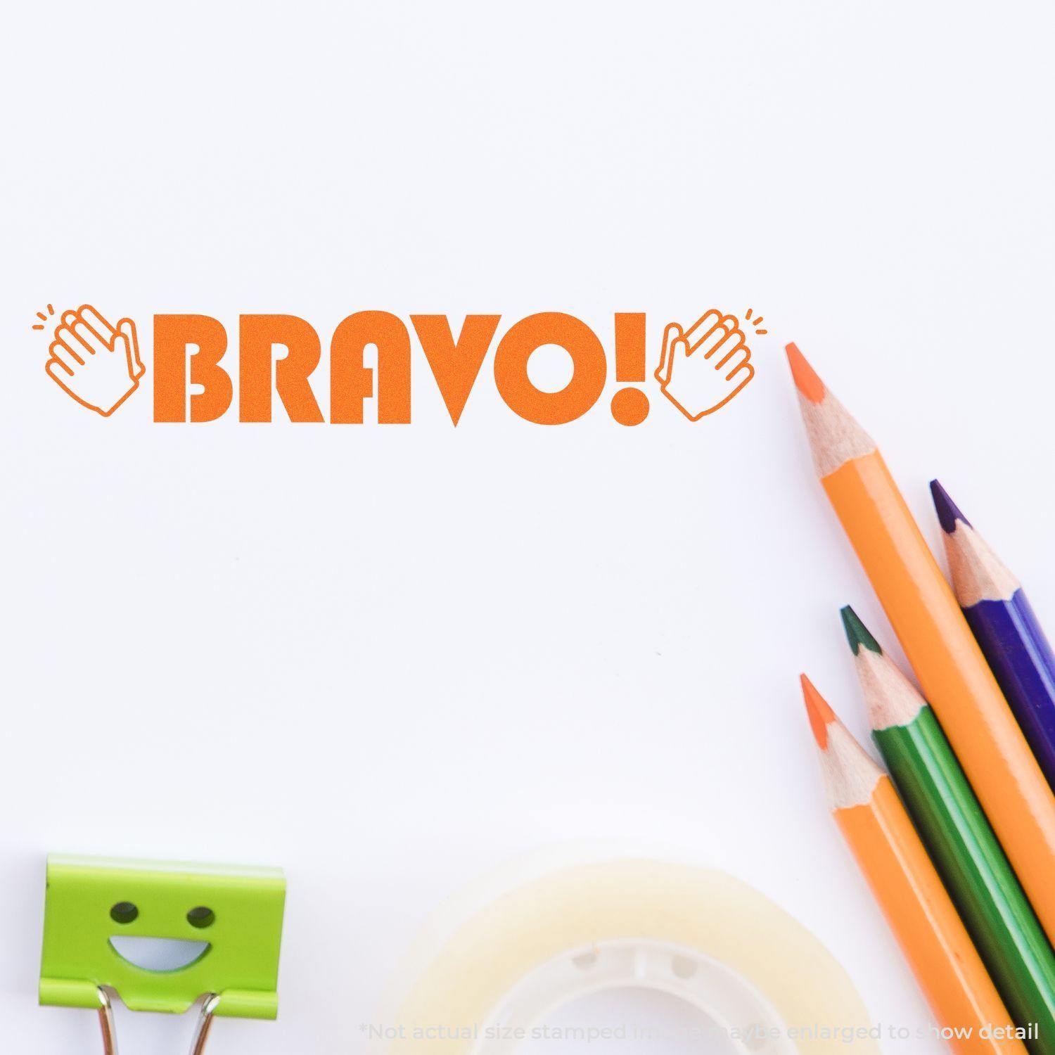 Large Bravo with Hands Rubber Stamp in orange ink on white paper, surrounded by colored pencils, a smiley face clip, and tape.