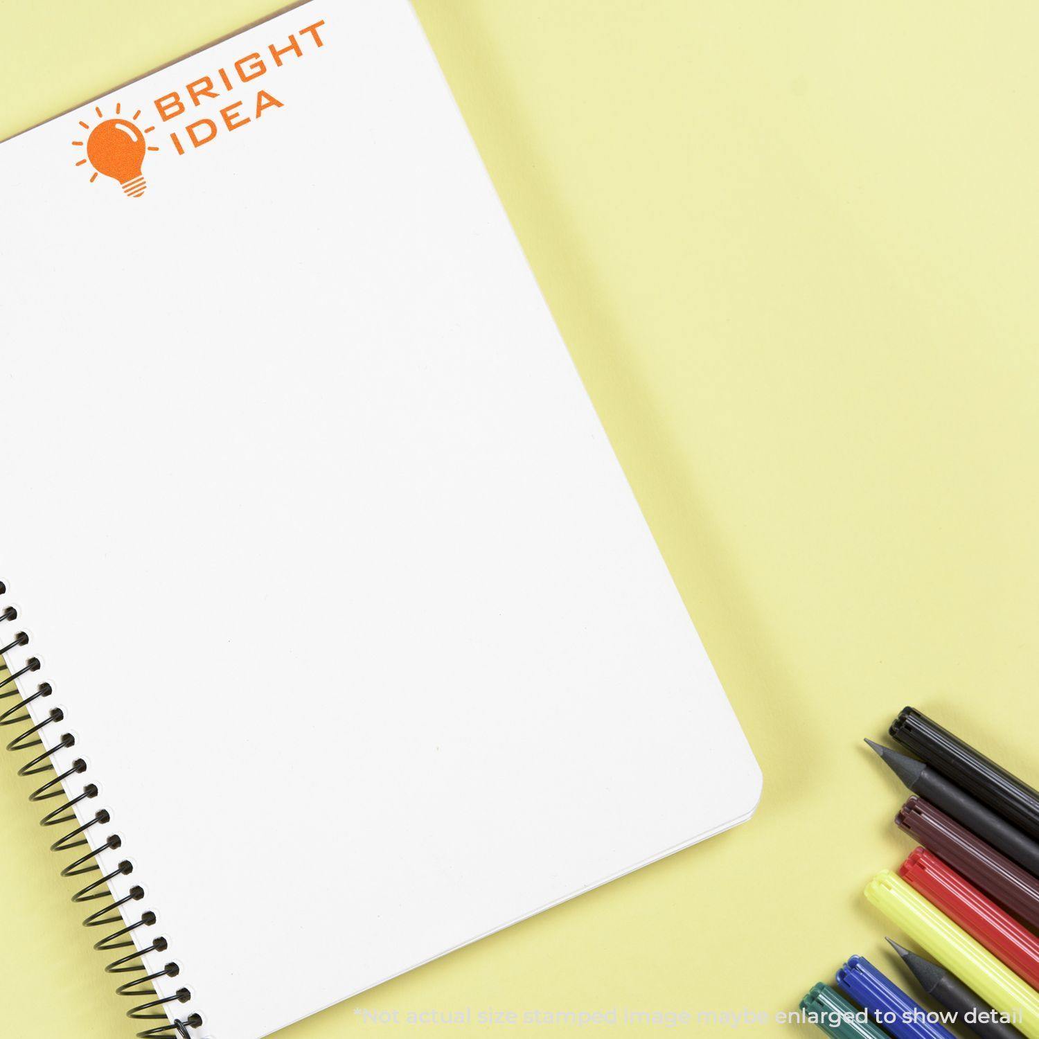 Self Inking Bright Idea Stamp imprint on a spiral notebook, surrounded by colorful pens on a yellow background.