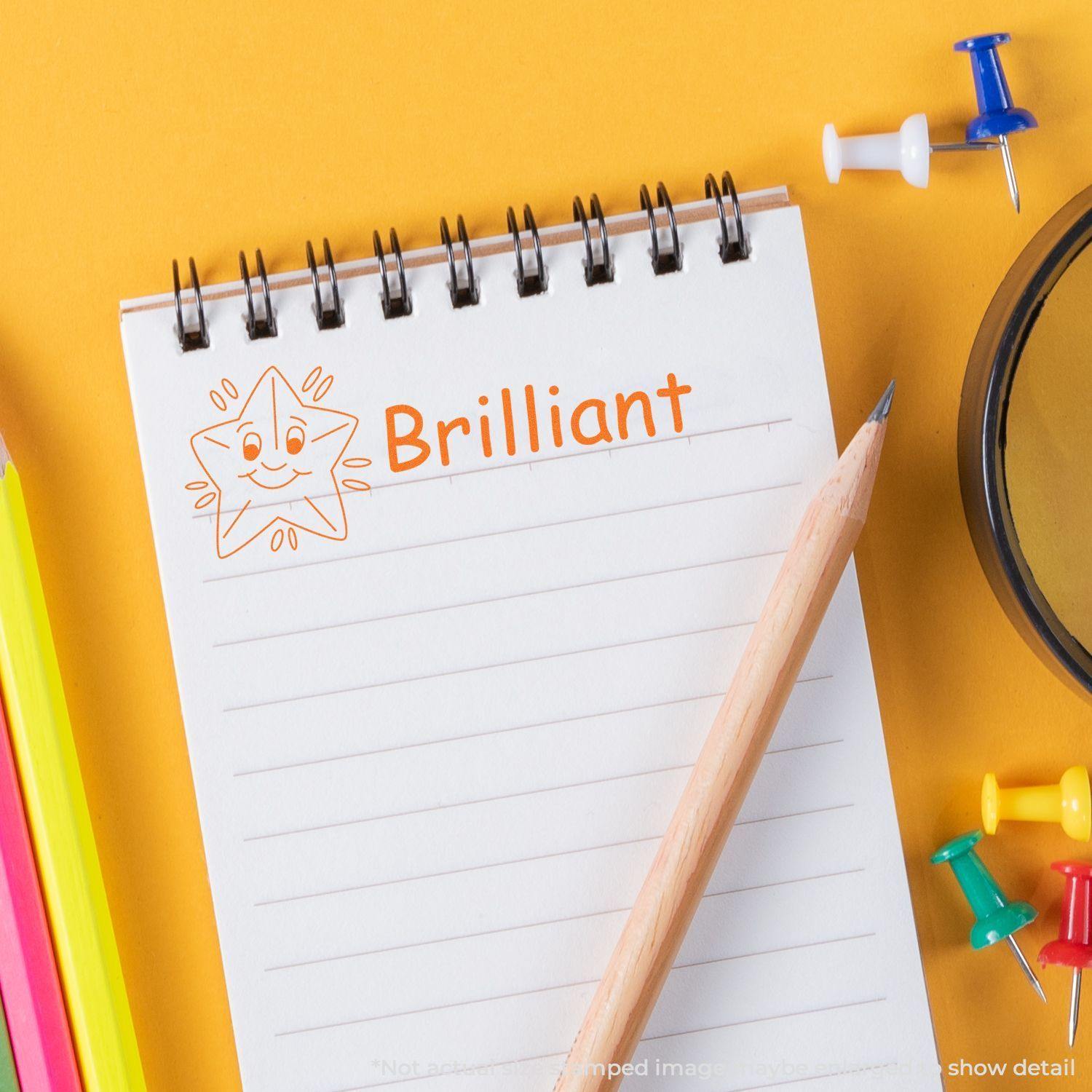 A notebook with a stamped image of a smiling star and the word Brilliant in orange ink, created using the Large Self Inking Brilliant Stamp. The notebook is surrounded by colorful pencils, push pins, and a pencil.