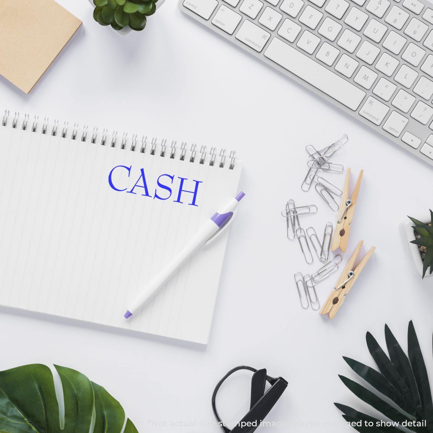 Desk with Slim Pre-Inked Cash Stamp on notebook, surrounded by keyboard, plants, pen, paper clips, and clothespins.