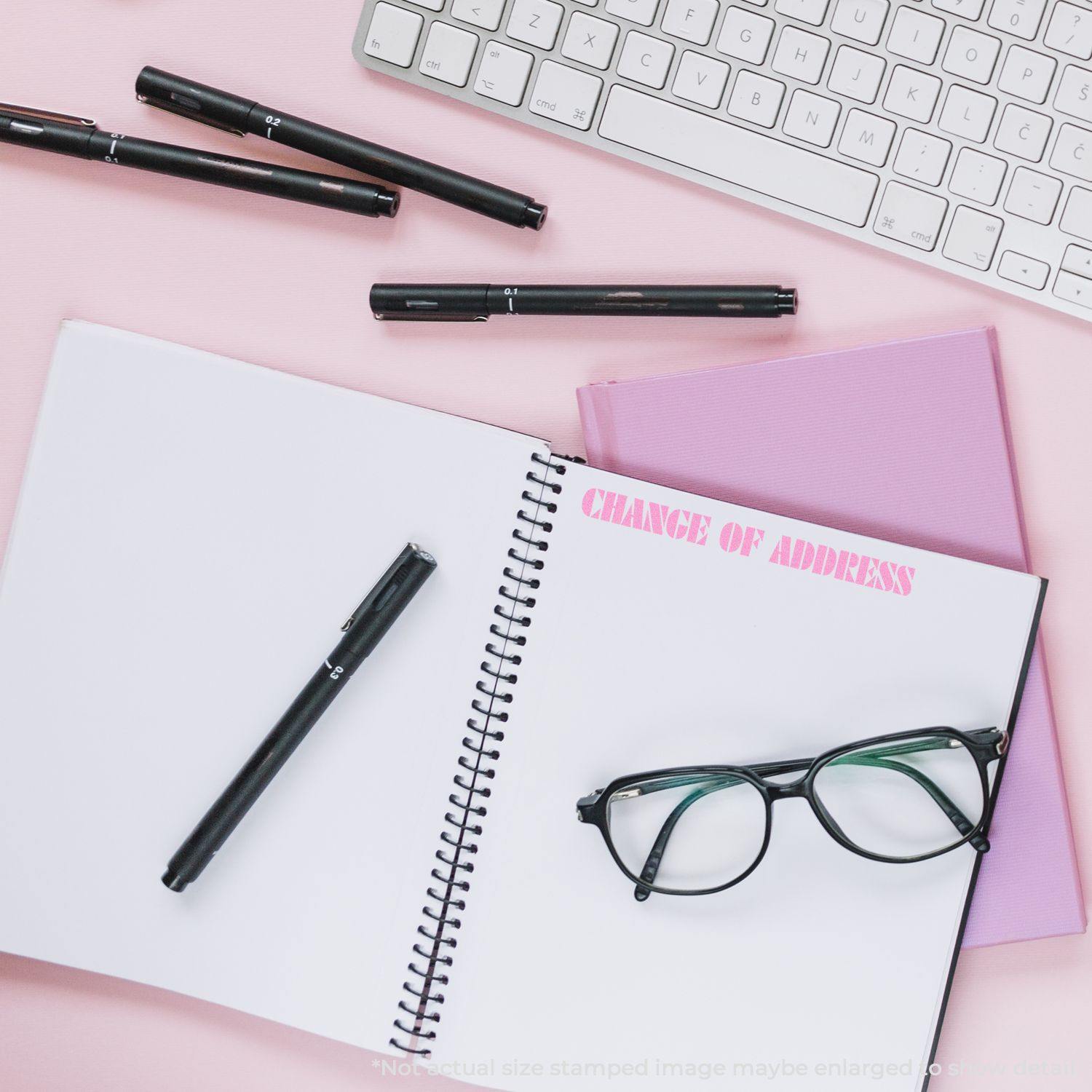 A Large Self Inking Change Of Address Stamp is shown in use on a spiral notebook with CHANGE OF ADDRESS stamped in pink ink. The scene includes a keyboard, pens, glasses, and a pink background.