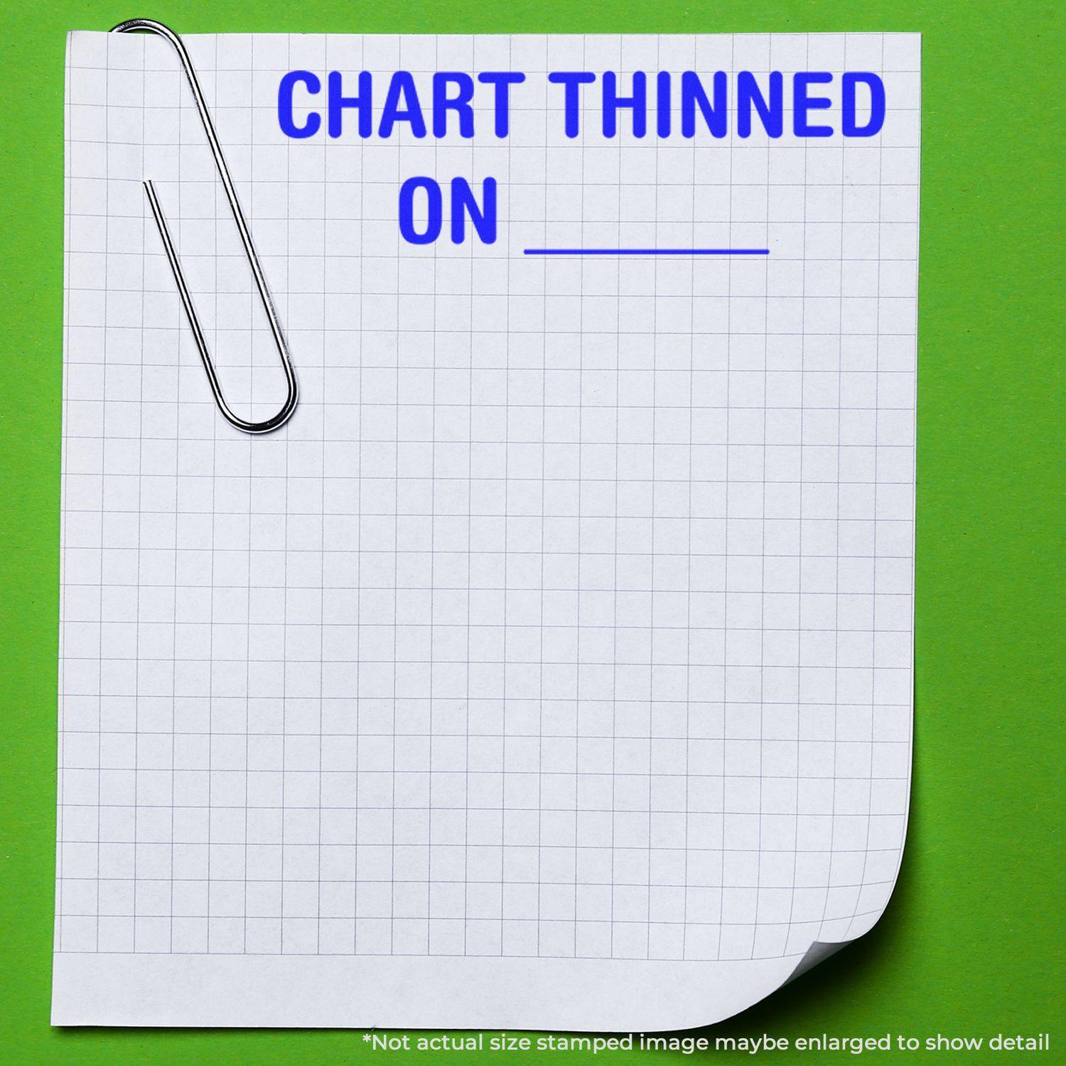 A piece of graph paper with a paperclip and the text CHART THINNED ON stamped in blue using the Large Self Inking Chart Thinned On Stamp. The paper is placed on a green background.