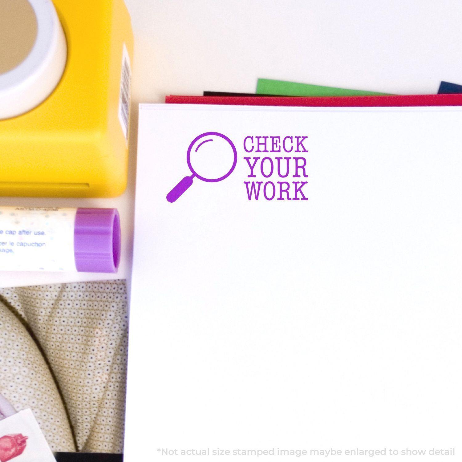 Self Inking Check Your Work Stamp in purple ink on a white paper, surrounded by office supplies including a yellow container and glue stick.