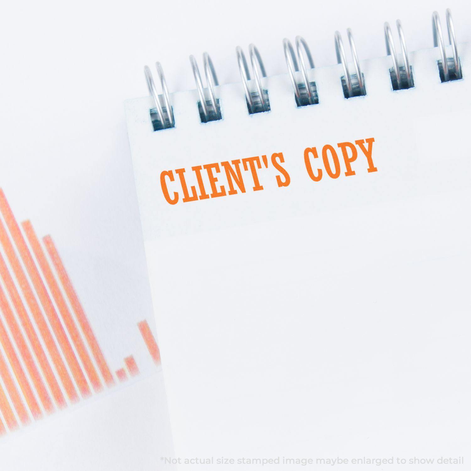 Large Self Inking Clients Copy Stamp imprinting 'CLIENT'S COPY' in orange ink on a white spiral notebook page, with a partial view of an orange bar graph in the background.