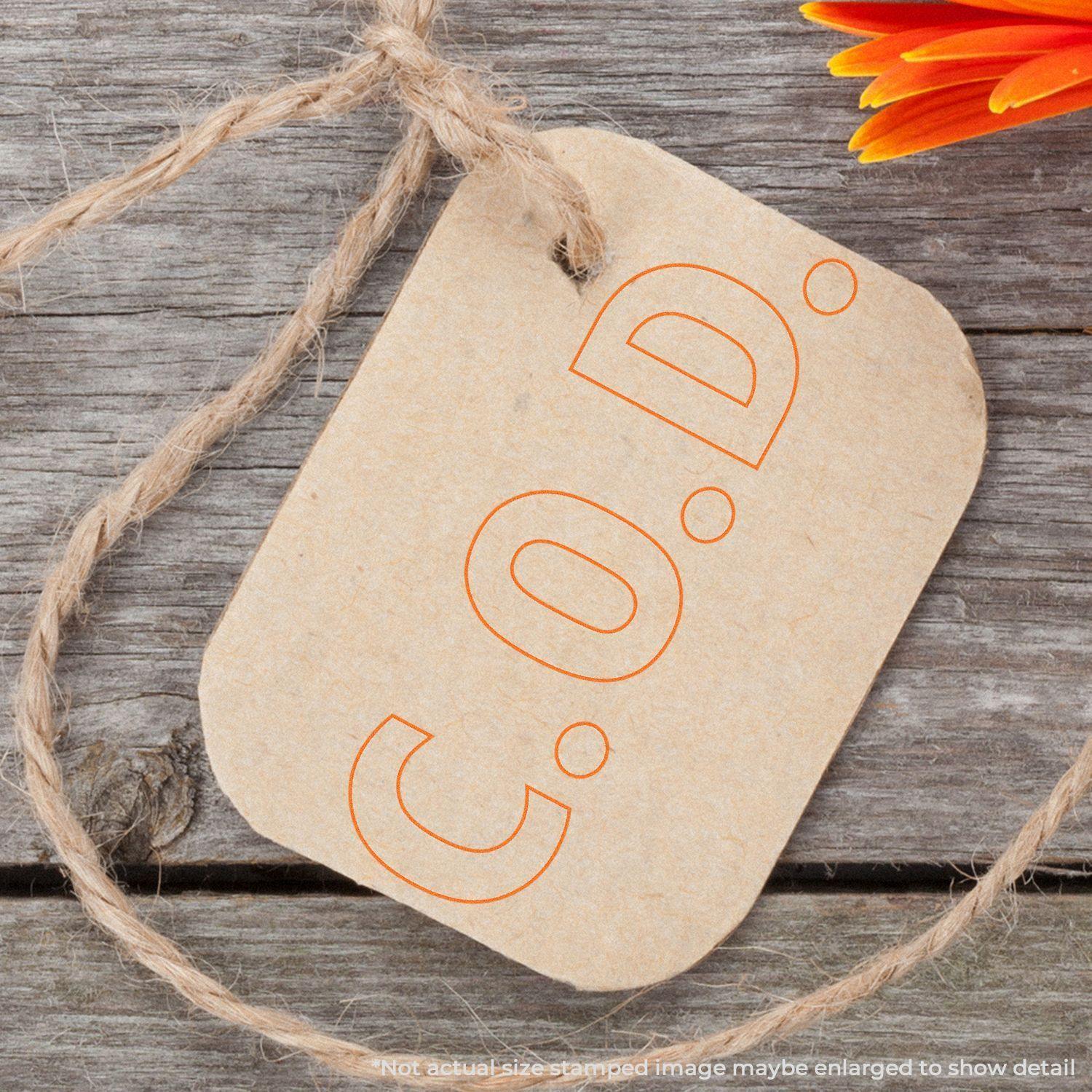 Slim Pre-Inked COD Stamp With Outline Text on a brown tag tied with twine, placed on a wooden surface with an orange flower.