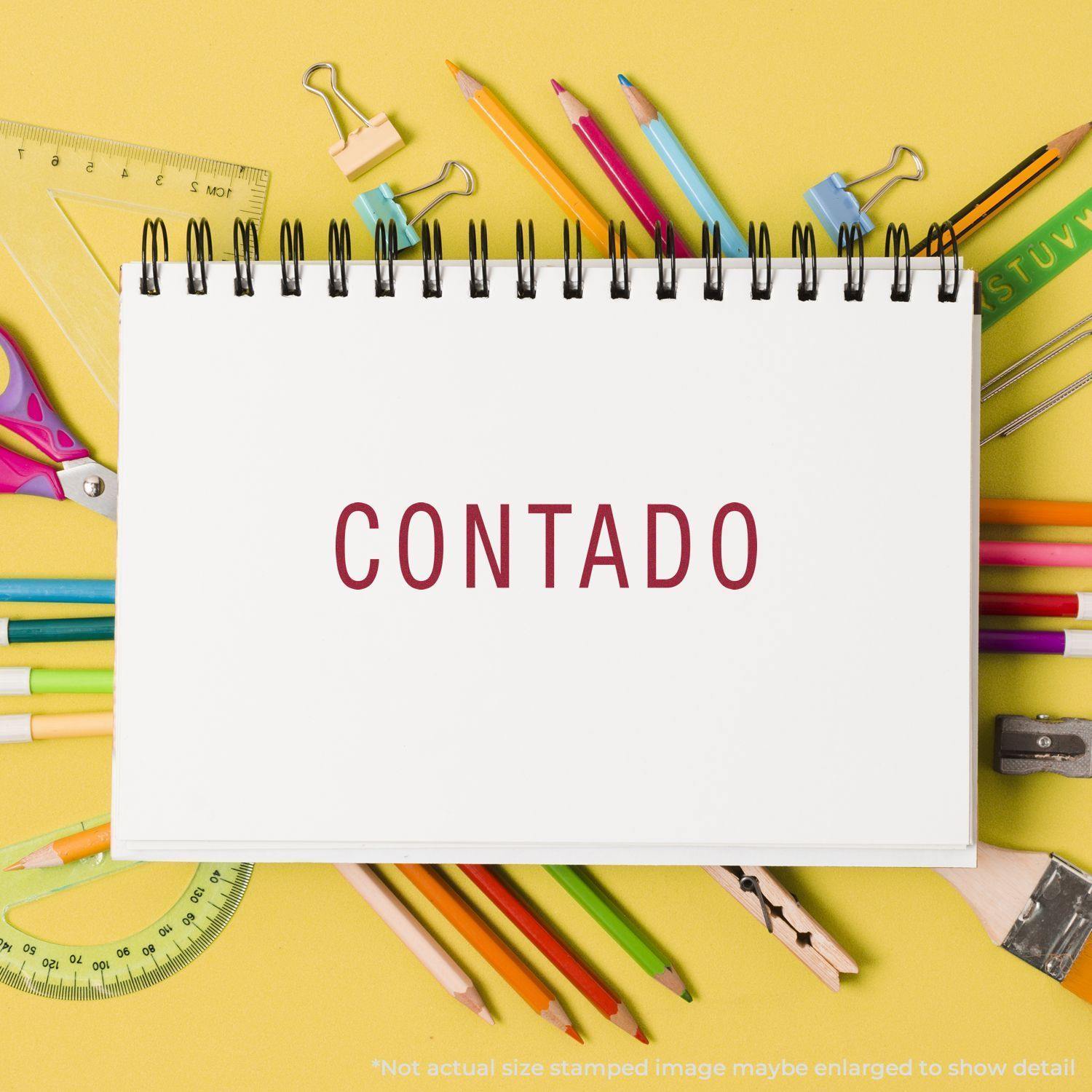 A Contado Rubber Stamp imprint on a white notepad surrounded by colorful stationery items on a yellow background.