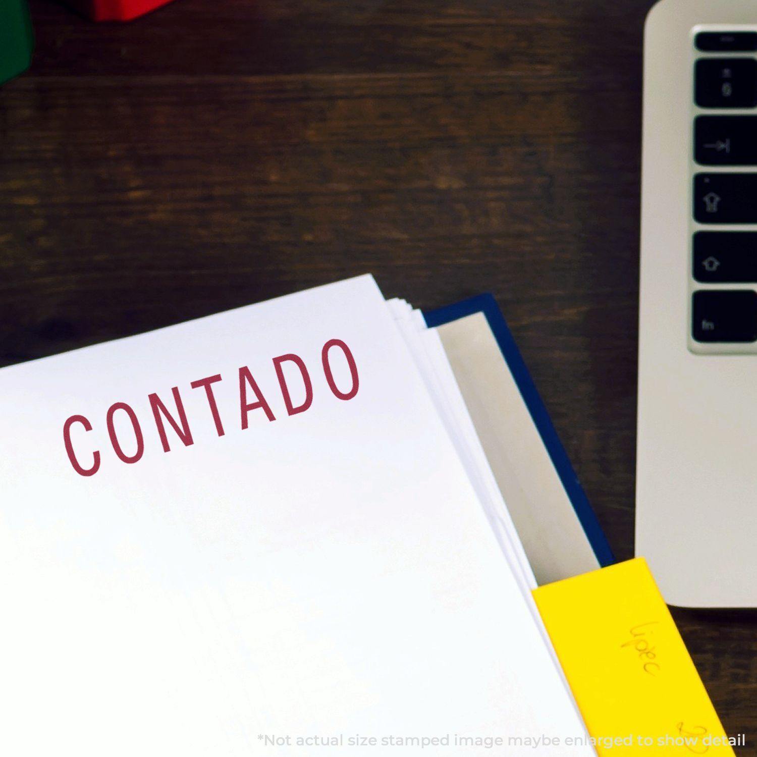 Large Self Inking Contado Stamp used on a white sheet of paper, leaving a clear red 'CONTADO' imprint. The paper is on a wooden desk next to a laptop and a yellow sticky note.