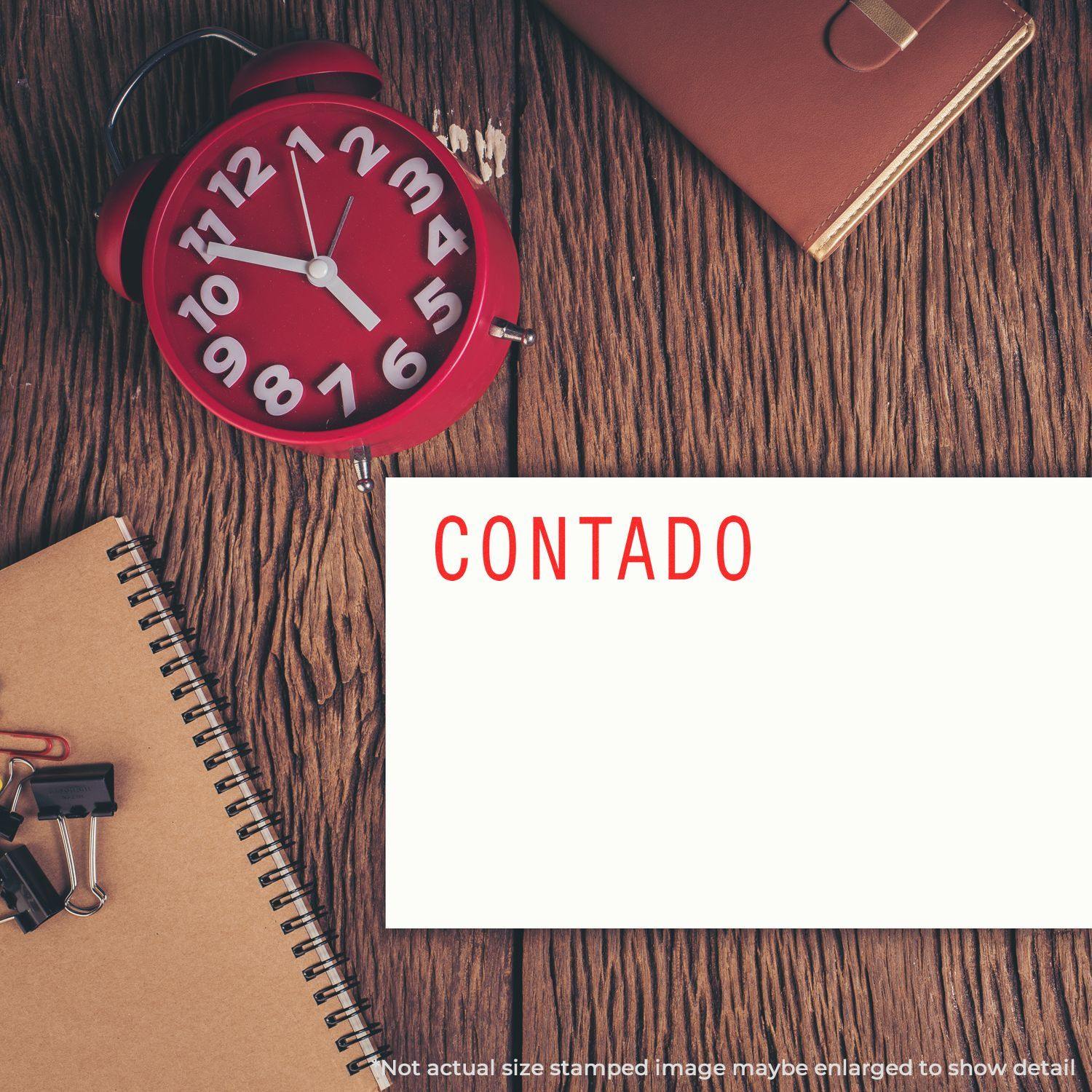 Large Pre-Inked Contado Stamp in use on a white card, placed on a wooden desk with a red clock, notebook, and office supplies.