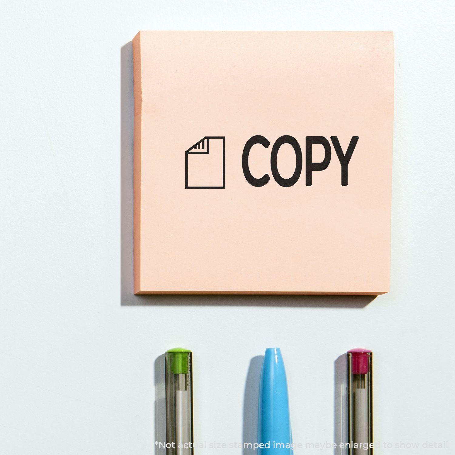 Large Self Inking Copy with Letter Stamp used on a pink sticky note, with three pens below it on a white surface.