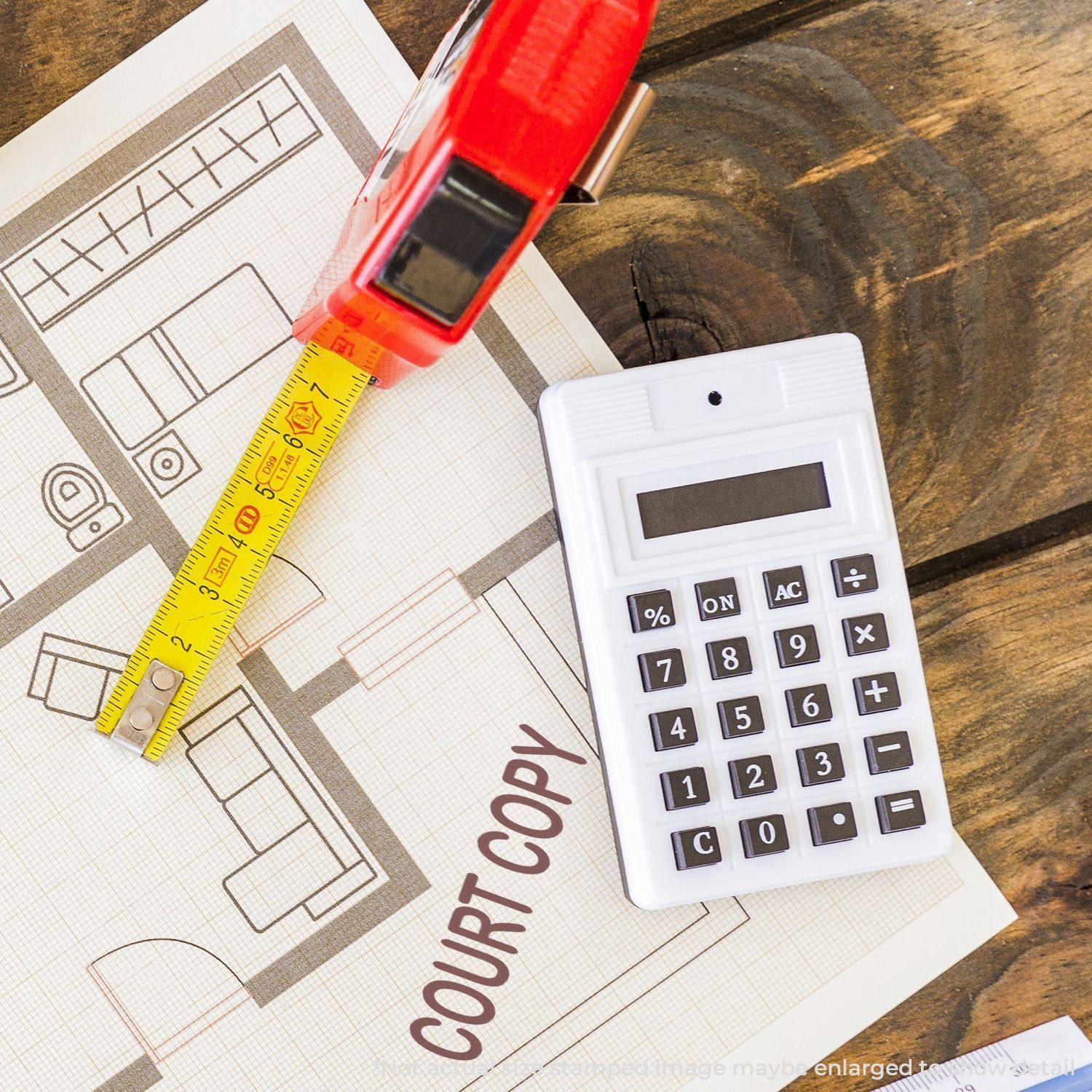 A Court Copy Rubber Stamp is placed on a floor plan next to a red tape measure and a white calculator on a wooden surface.