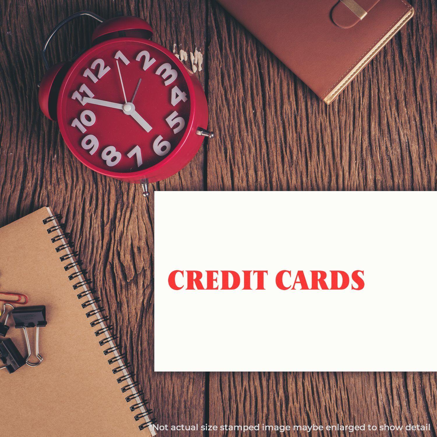 A Self Inking Credit Cards Stamp on a wooden desk with a red clock, notebook, binder clips, and a leather-bound book.