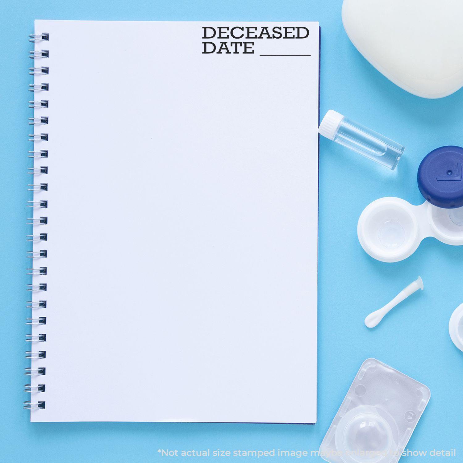 Large Self Inking Deceased Date Stamp used on a blank notebook page, surrounded by various small office and personal items on a blue surface.