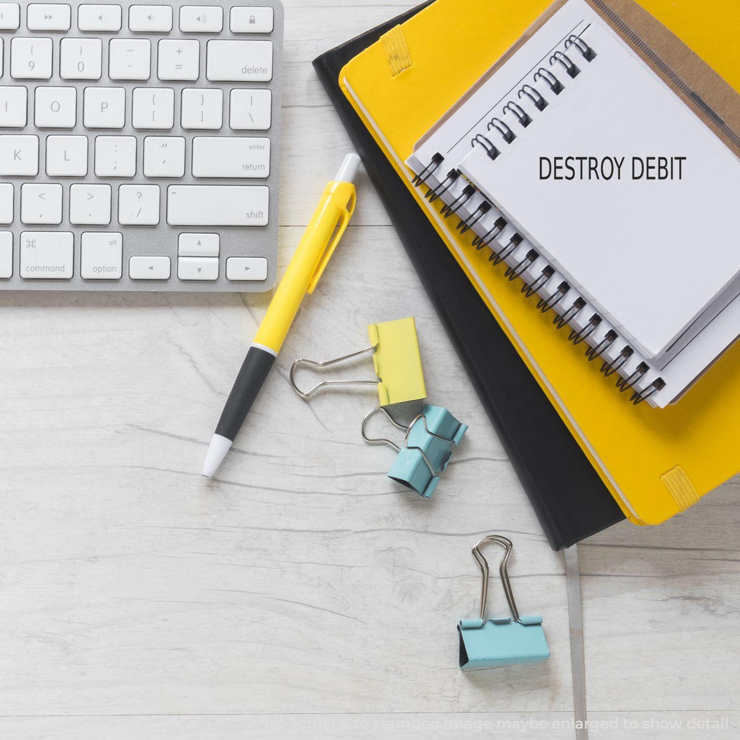 A Self Inking Destroy Debit Stamp is shown on a notepad next to a keyboard, yellow pen, and colorful binder clips on a desk.