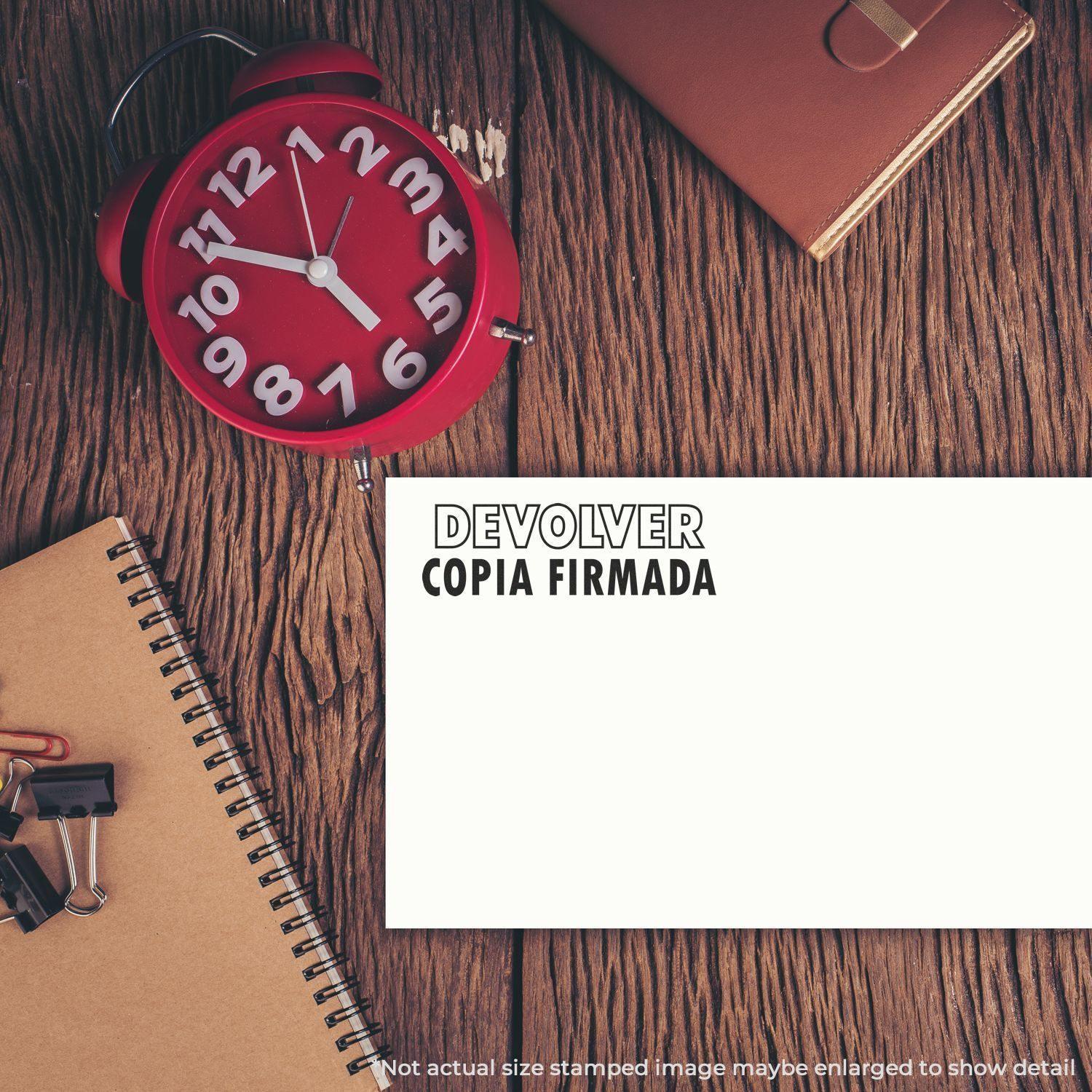 Desk with a red clock, notebook, and paper stamped with 'Devolver Copia Firmada' using a Slim Pre-Inked Devolver Copia Stamp.