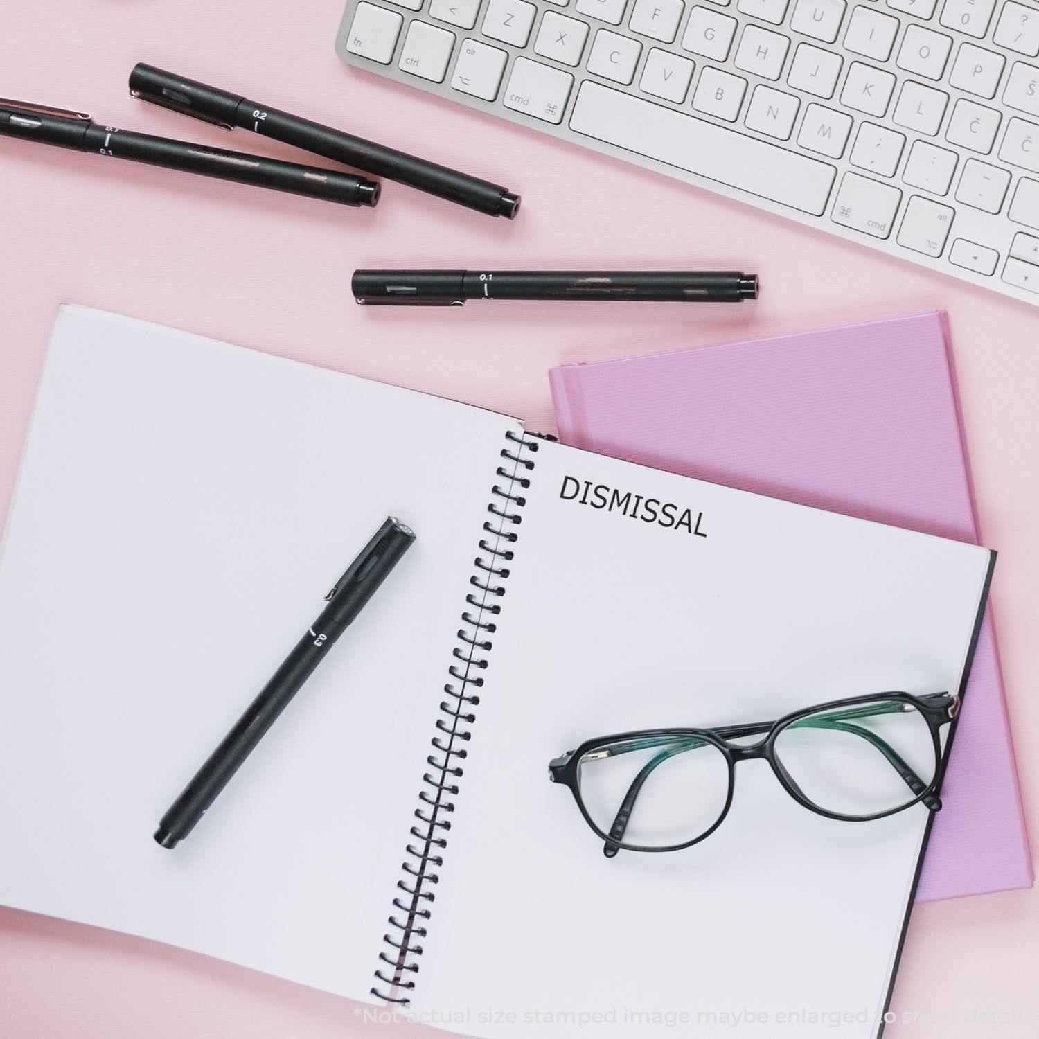 Large Dismissal Rubber Stamp used on a notebook, surrounded by pens, glasses, a keyboard, and a pink folder on a pink desk.