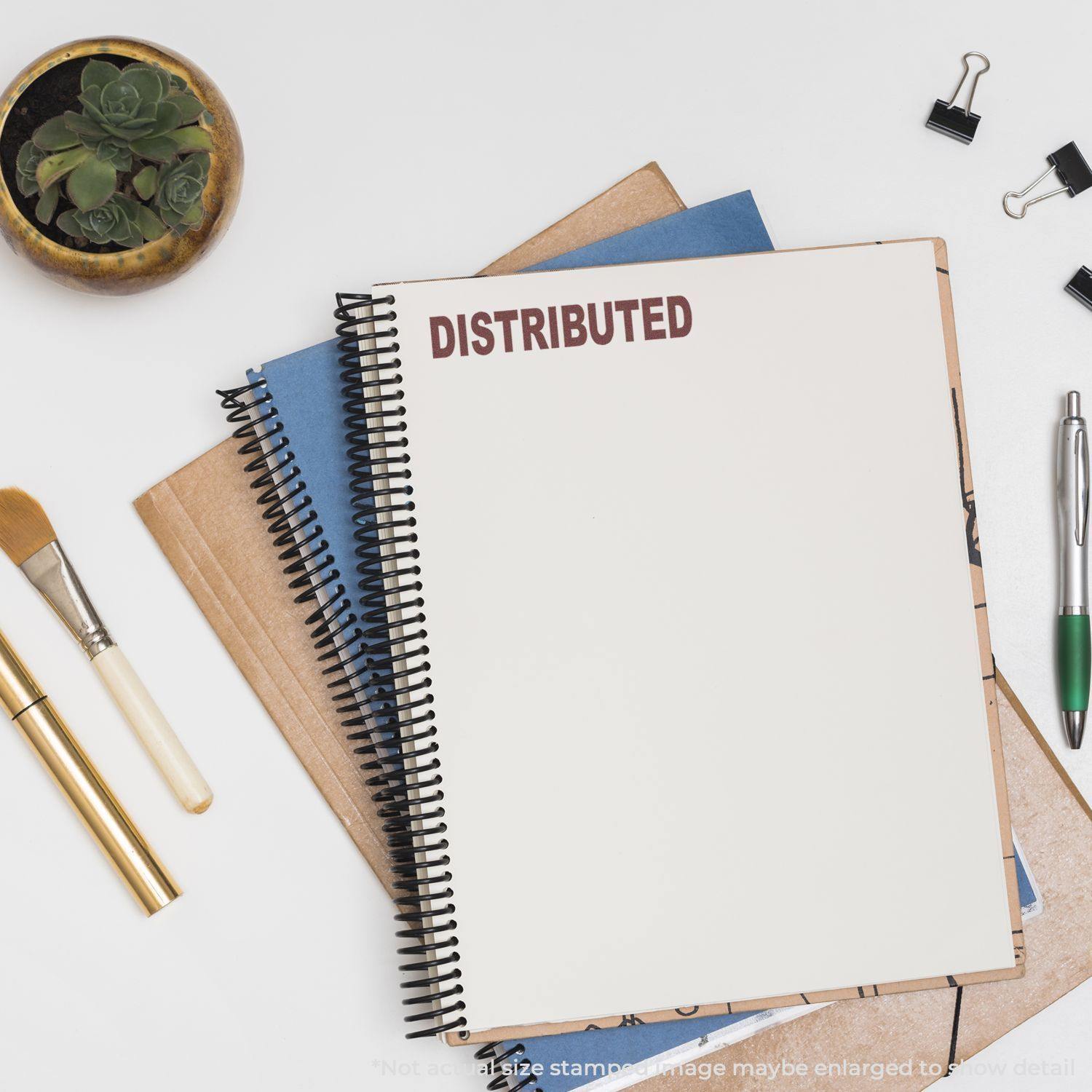 Self-Inking Distributed Stamp marking DISTRIBUTED on a spiral notebook, surrounded by office supplies including pens, clips, and a plant.