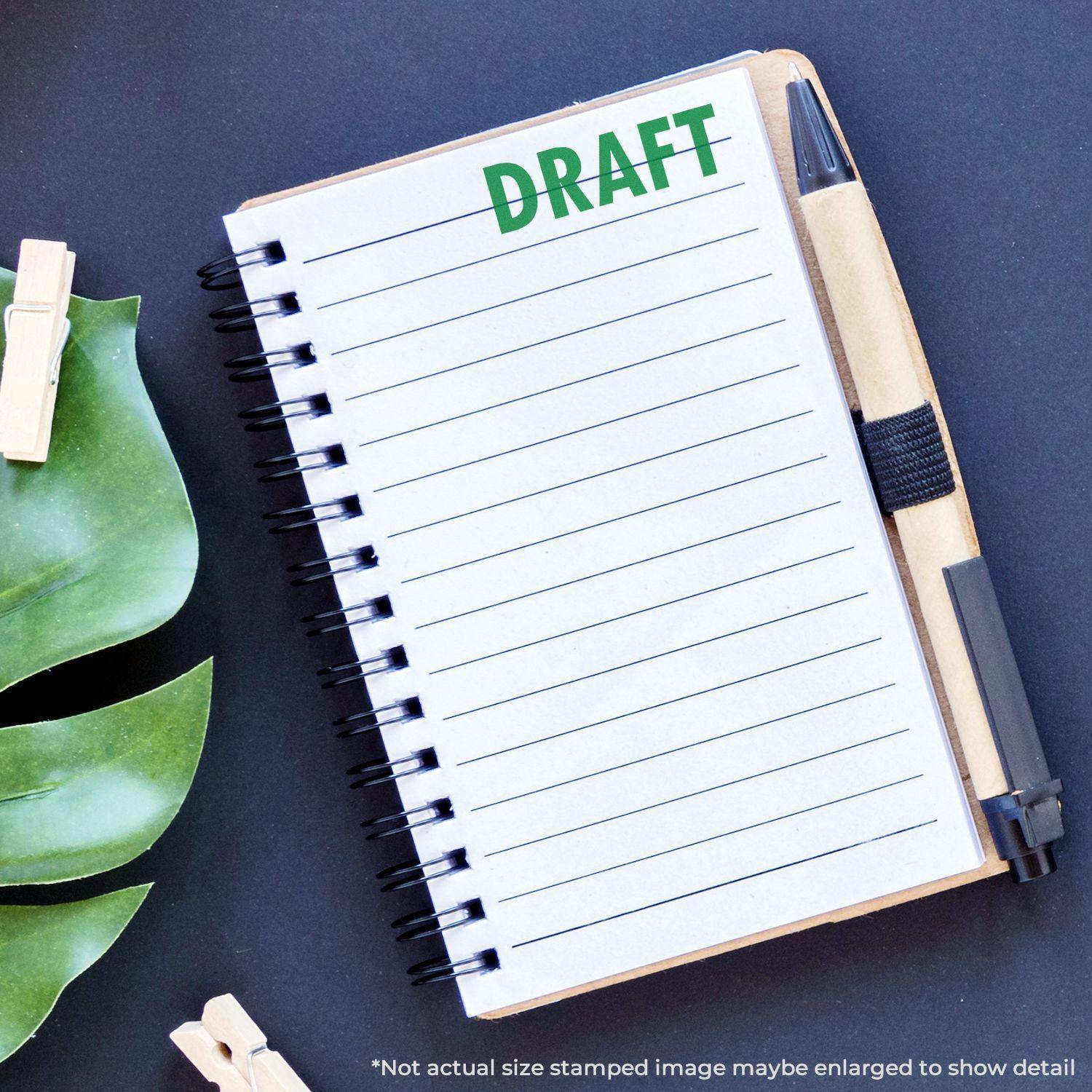 A notebook stamped with DRAFT using the Large Draft Rubber Stamp, placed on a desk with a pen, leaves, and clothespins.